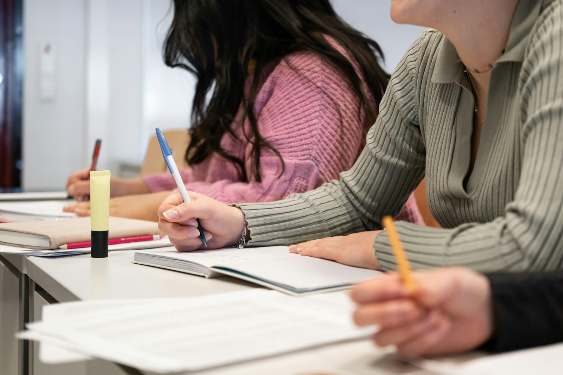 Three students are seated at a desk, engaged in note-taking with various pens and a highlighter. Their notebooks are open, suggesting active learning.