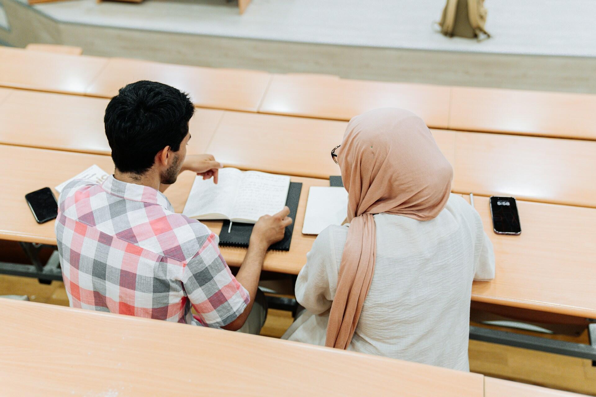 Two students studying together with books on a wooden lecture hall desk.