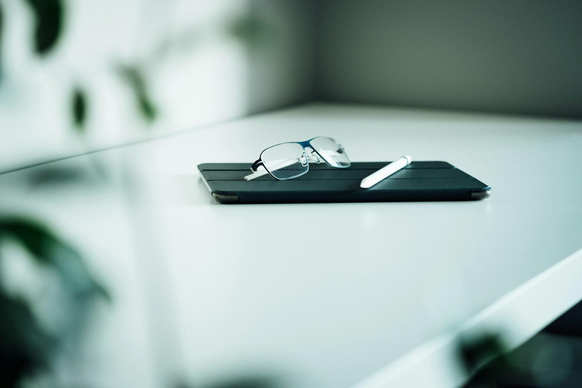Eyeglasses on a tablet with a stylus on a white desk.