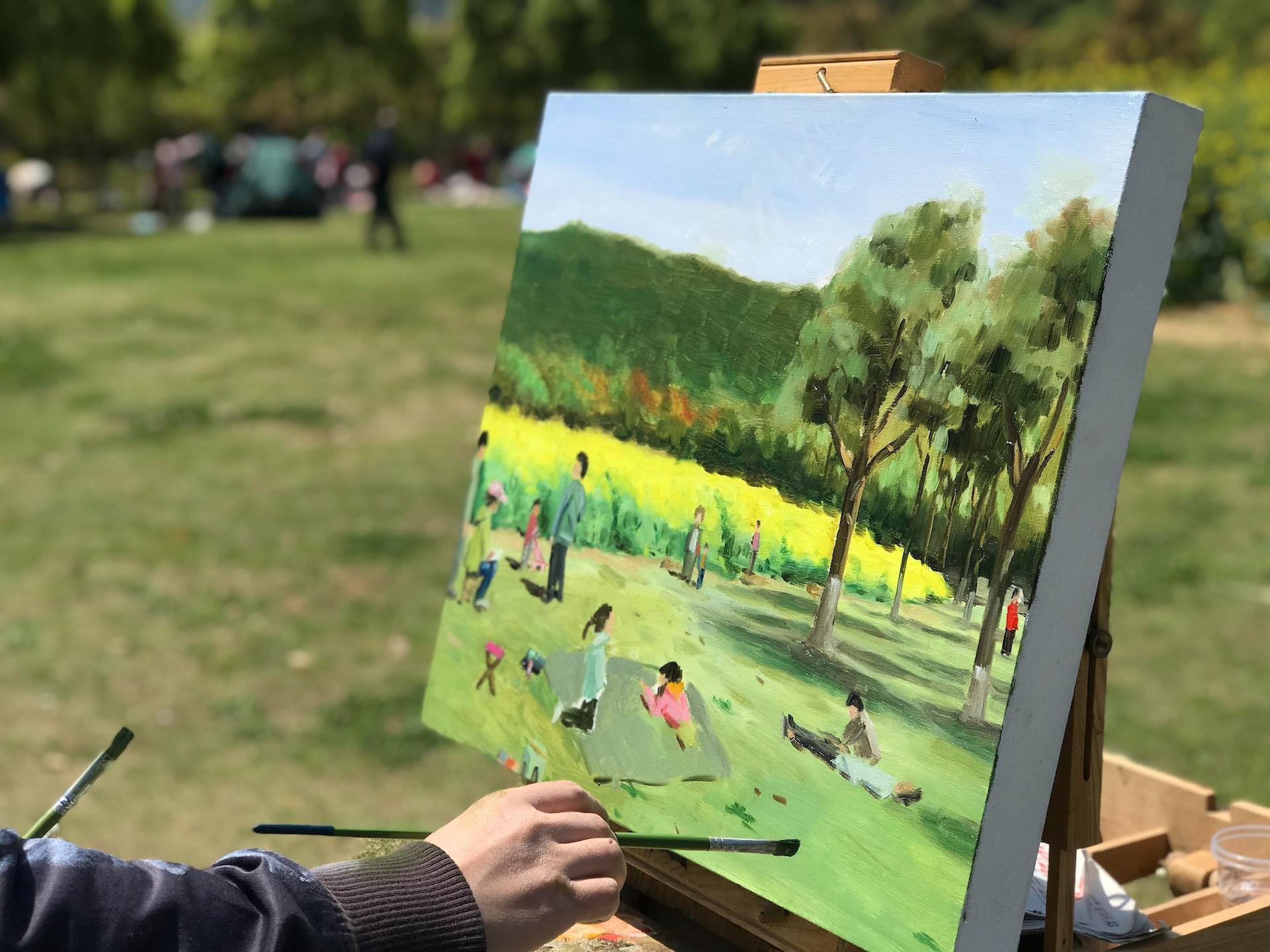 An artist wearing a long-sleeved dark shirt and holding a paintbrush in his hand paints a meadow scene while standing in the sunny meadow. 