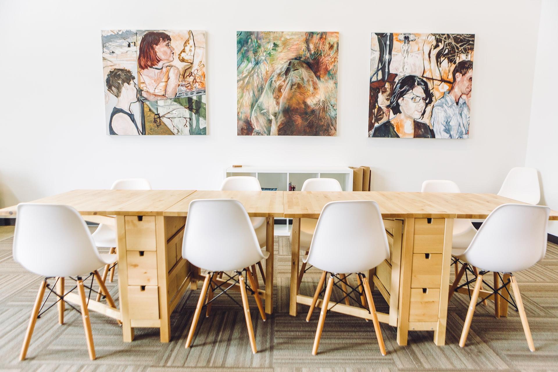 Three square canvasses depicting various people painted in brown tones hanging above a wooden table with two white chairs in front of it. 