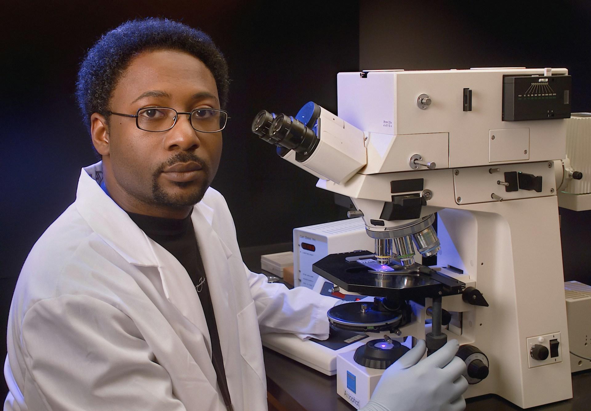 A man in lab white coat sitting next to a microscope.