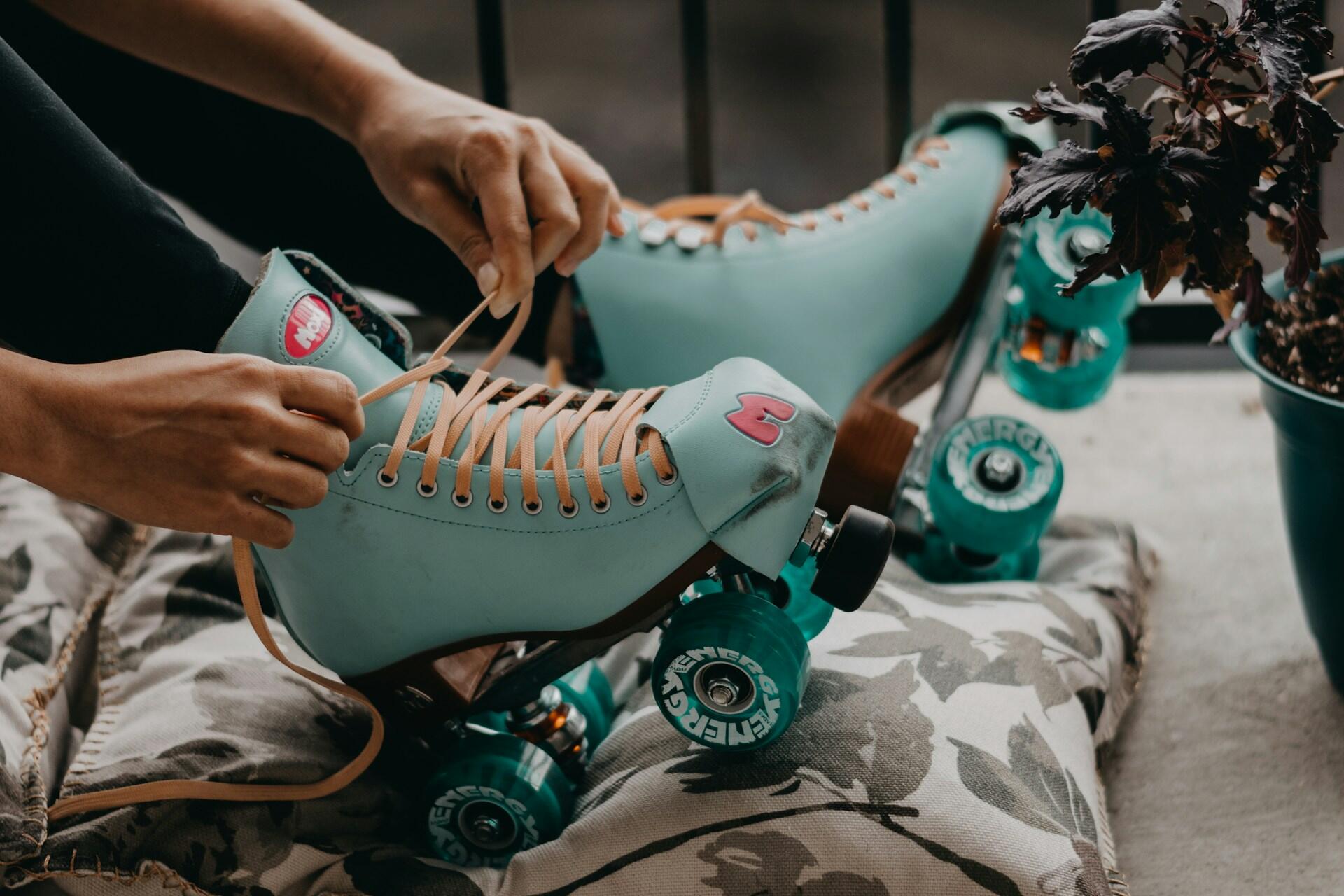 A close-up of a person lacing up light blue roller skates with teal wheels, set against a floral pillow and a potted plant.