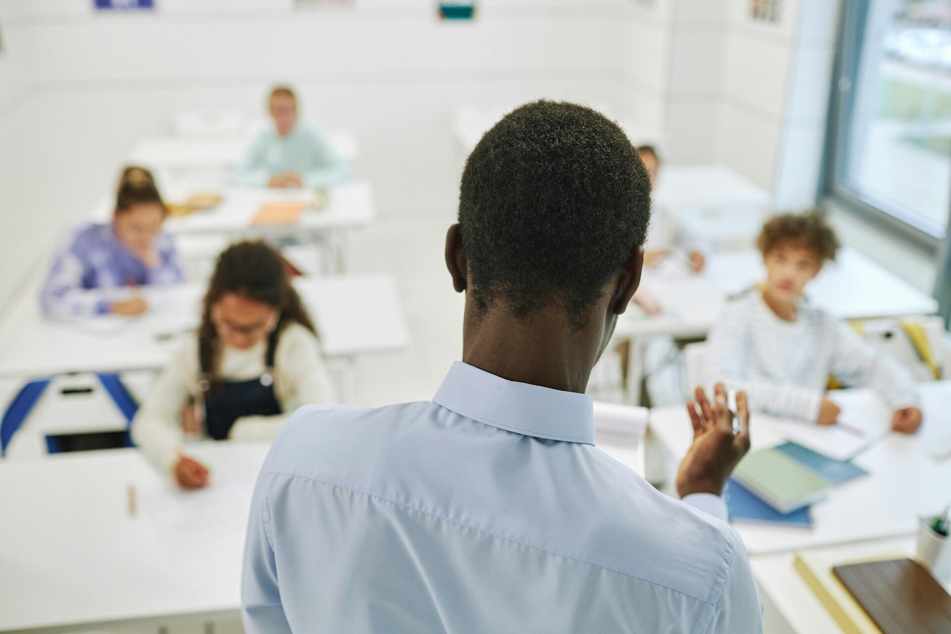 A teacher stands in front of students seated at desks, engaging in a lesson.