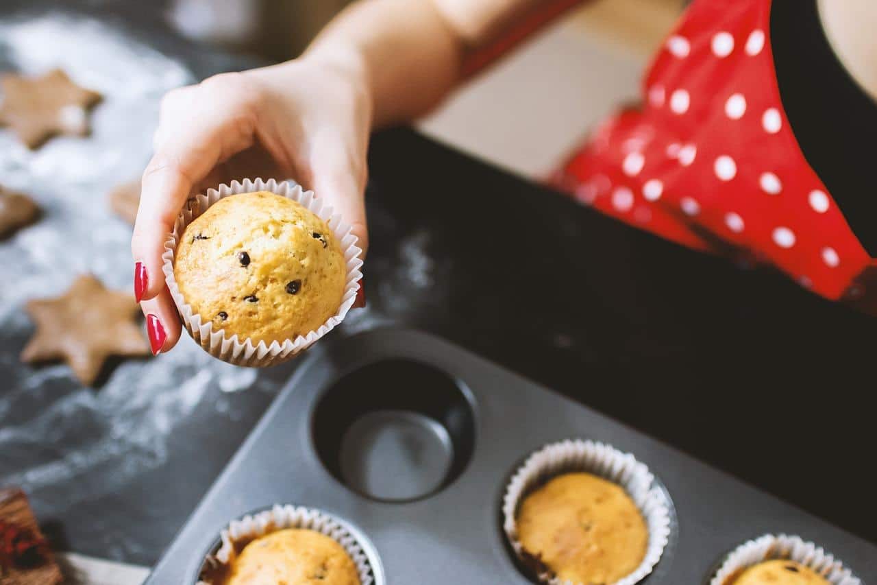 A person wearing a red polka-dot apron over a black top holds a cupcake in their right hand with its red-painted fingernails. A baking tin of similar muffins lays on a black surface next to a floured spot featuring star-shaped biscuits.