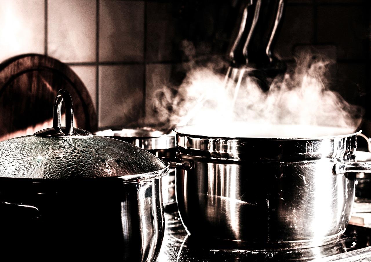 Two stainless steel pots atop a cooking surface. One has a glass lid showing condensation beads while the other exudes steam from its simmering contents. The white tile backdrop is mostly in shadow, appearing more sepia-toned.