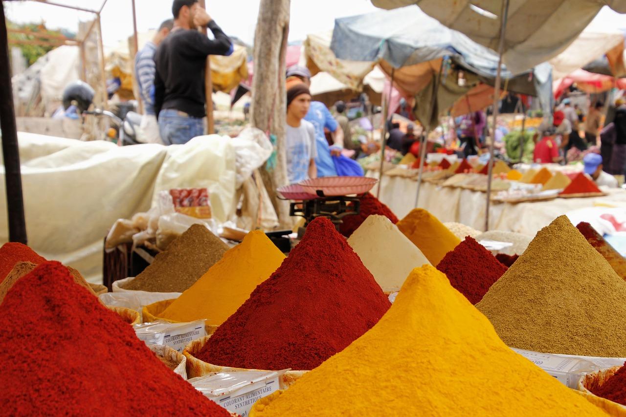 An open-air spice market featuring heaped barrels of red, yellow and beige spices. In the background, a vendor wearing a black top and denims stands behind a youth wearing a white top with a light blue pattern on it.