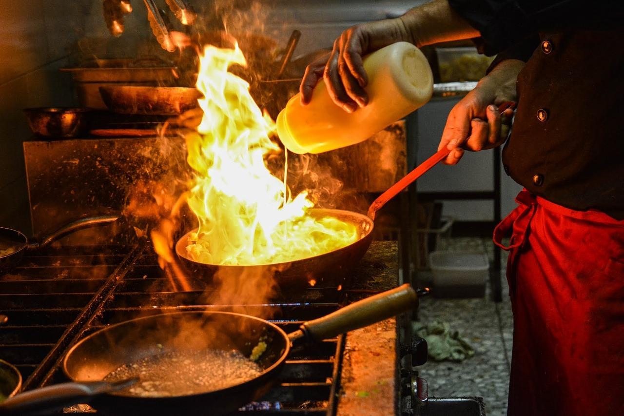 A person in a dimly-let setting, adding oil and agitating a wok from which flames shoot out.