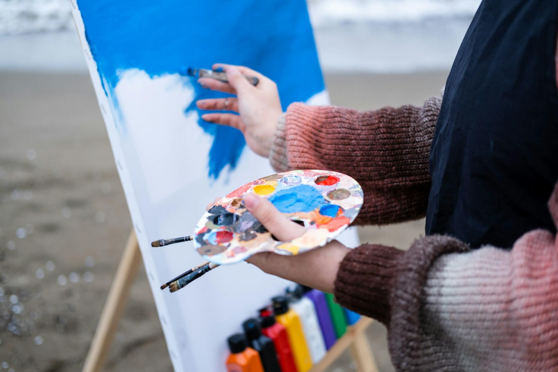 Person painting a blue sky on a canvas at the beach, holding a palette with various paint colours and brushes.