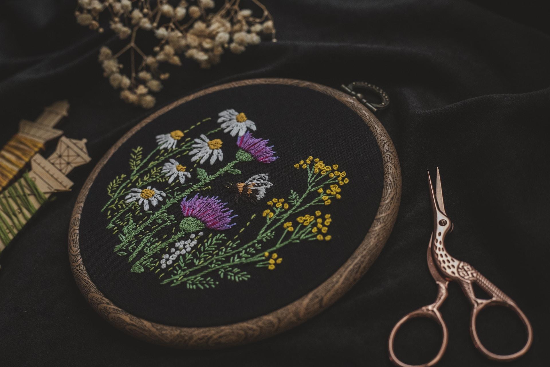 A wooden embroidery hoop encircles a field of embroidered flowers blooming on a black cloth.