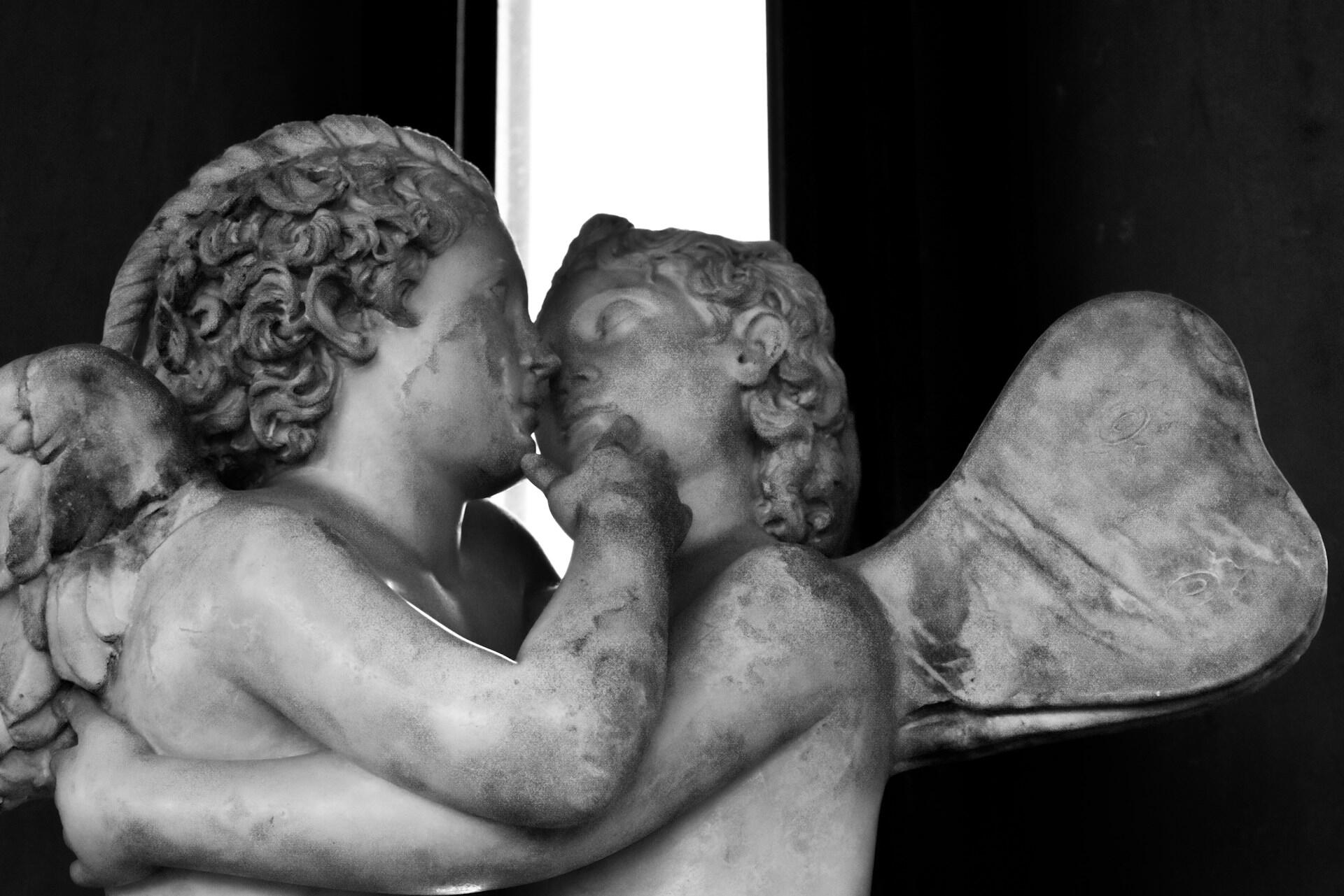 Two cherub statues embrace, their detailed wings and curly hair highlighted in black and white, conveying a sense of intimacy and affection.