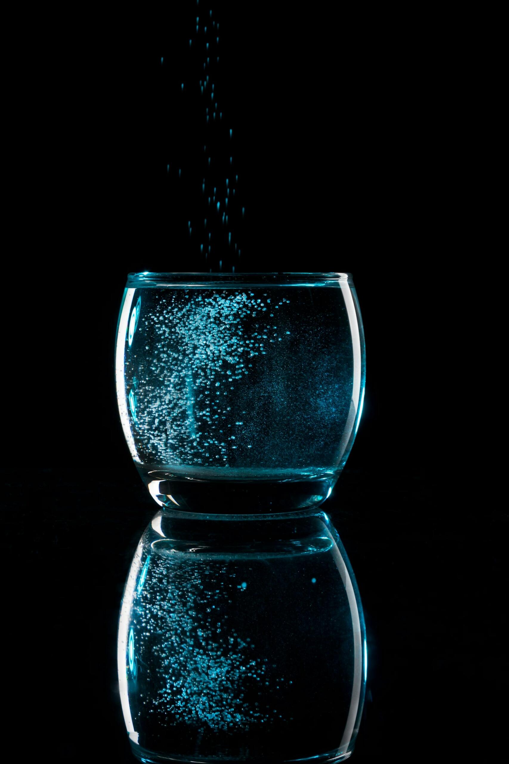 A glass of water with white crystals sprinkled into it against a black background.