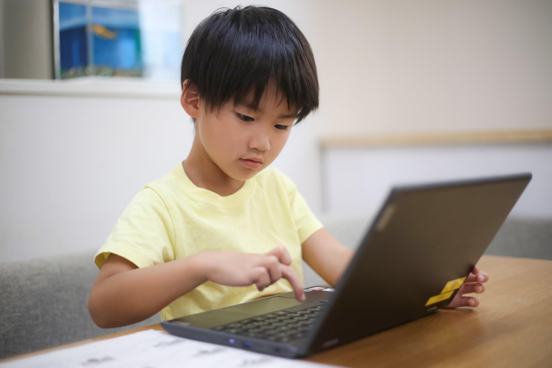 A boy wearing a yellow shirt uses his right hand to press a button on a laptop's keyboard while he stares at the screen.