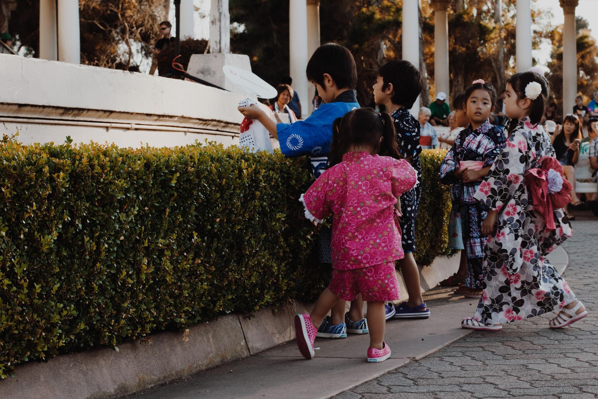 A group of boys wearing western dress and girls wearing flowered kimonos gather near a low hedge as parent, in the distance, look on.