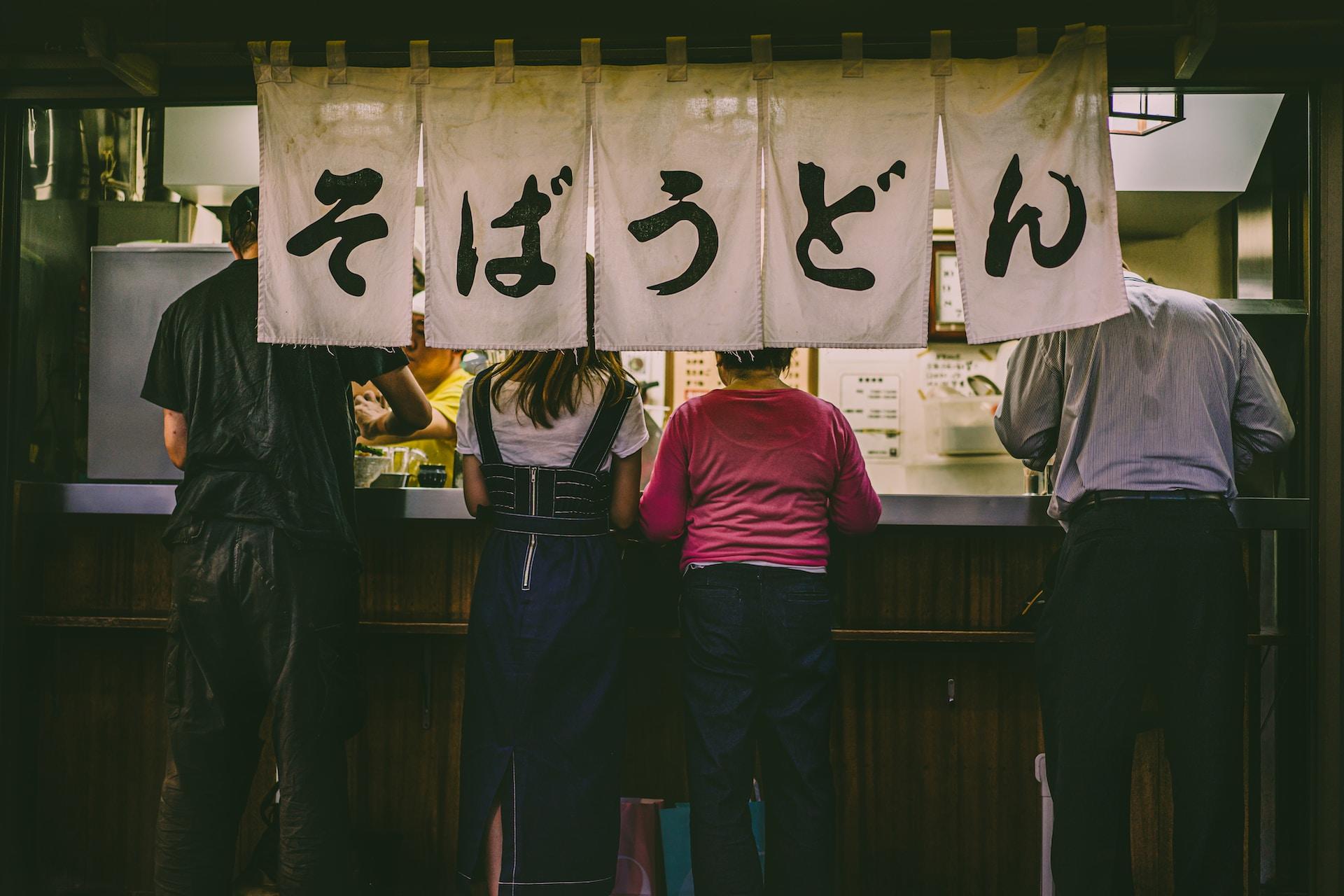 A food vendor stall open at night with several customers wearing cold weather clothes standing in front. Five sheets of white cloth, each with a Japanese pictogram painted on in black ink, hand from the overhang. 