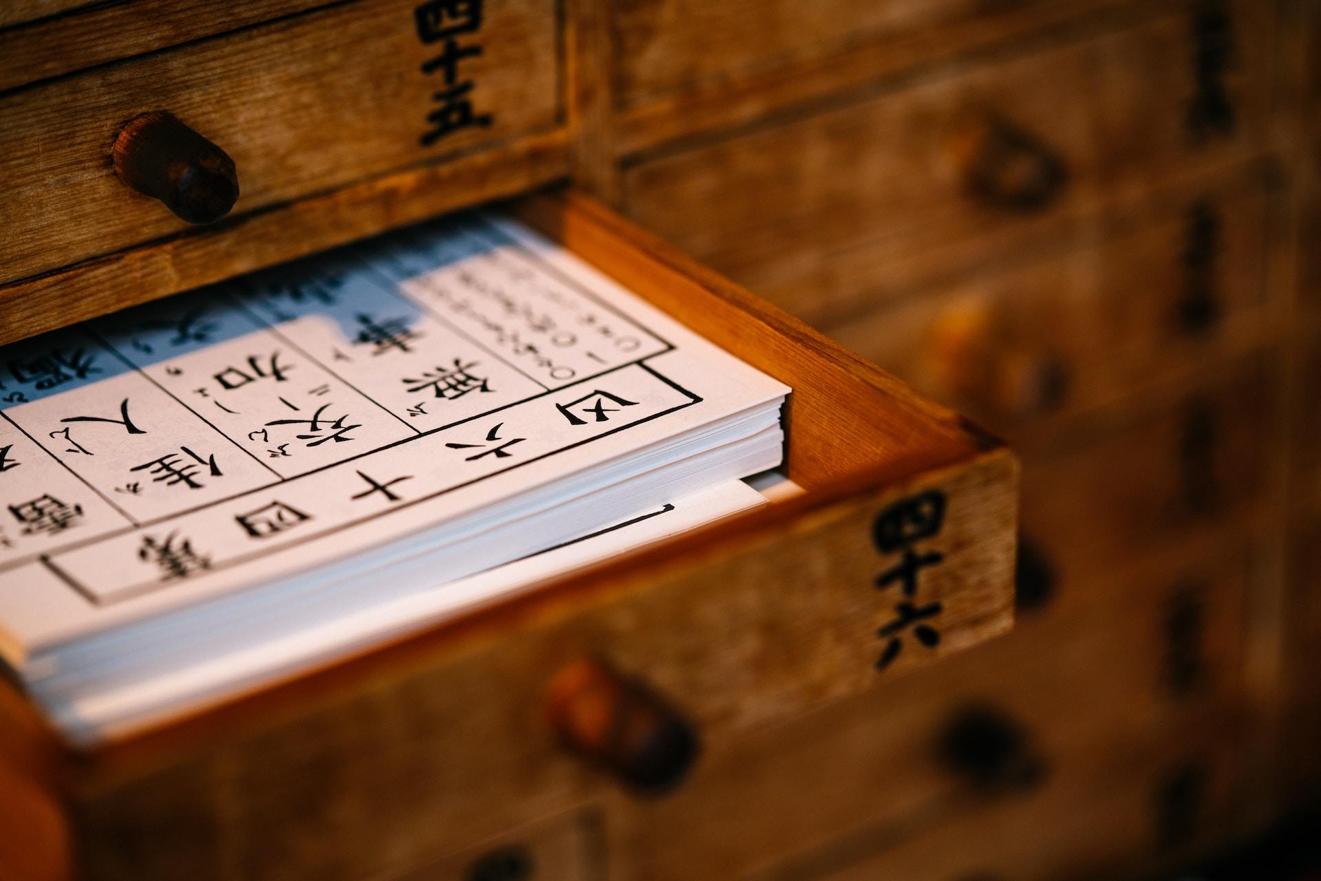 A drawer of a wooden cabinet opened to reveal white sheets of paper with black kanji pictograms written on them.