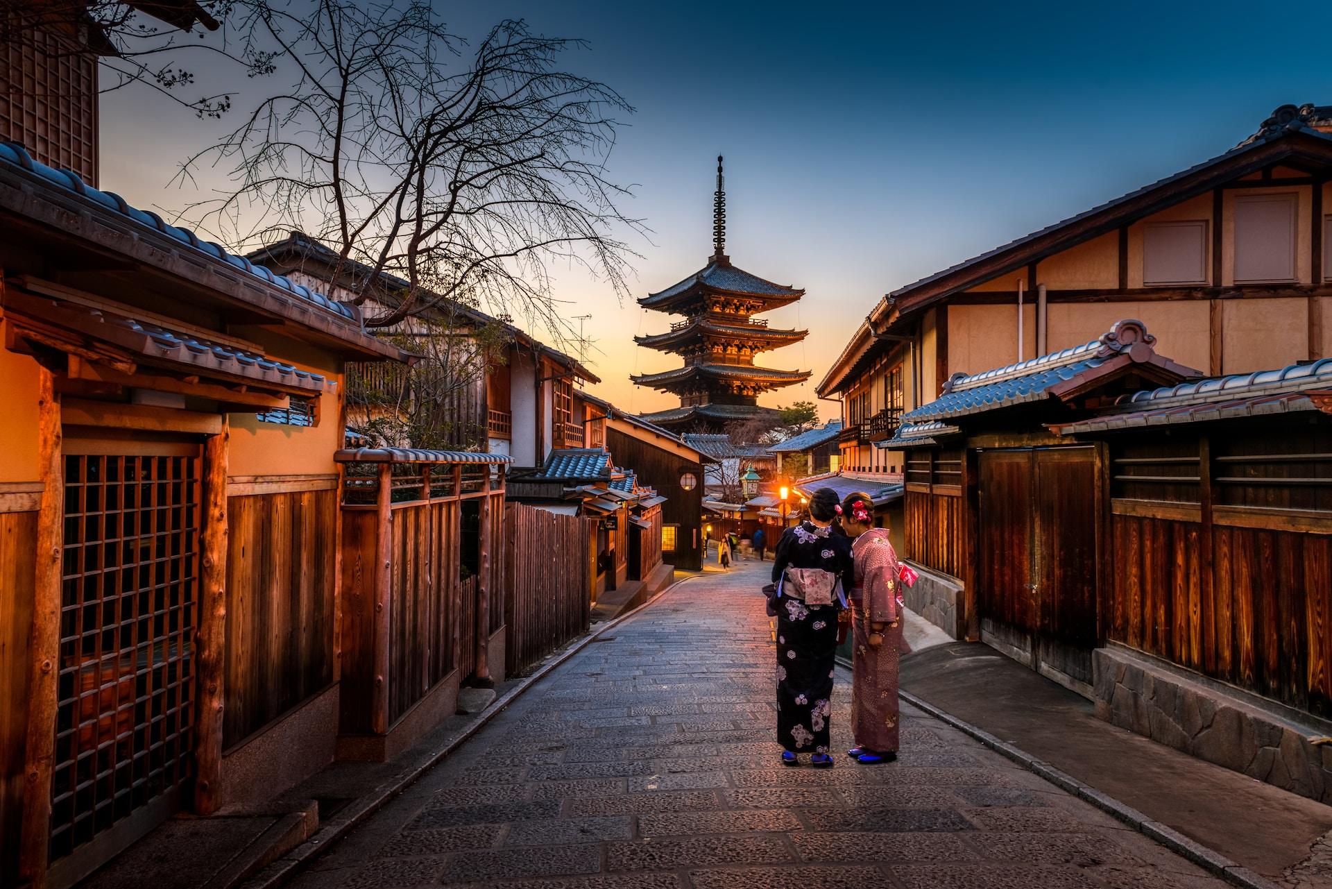 Two women wearing kimonos stand in a deserted alleyway at dawn, the walkway is flanked on both sides with wood-fronted low buildings.