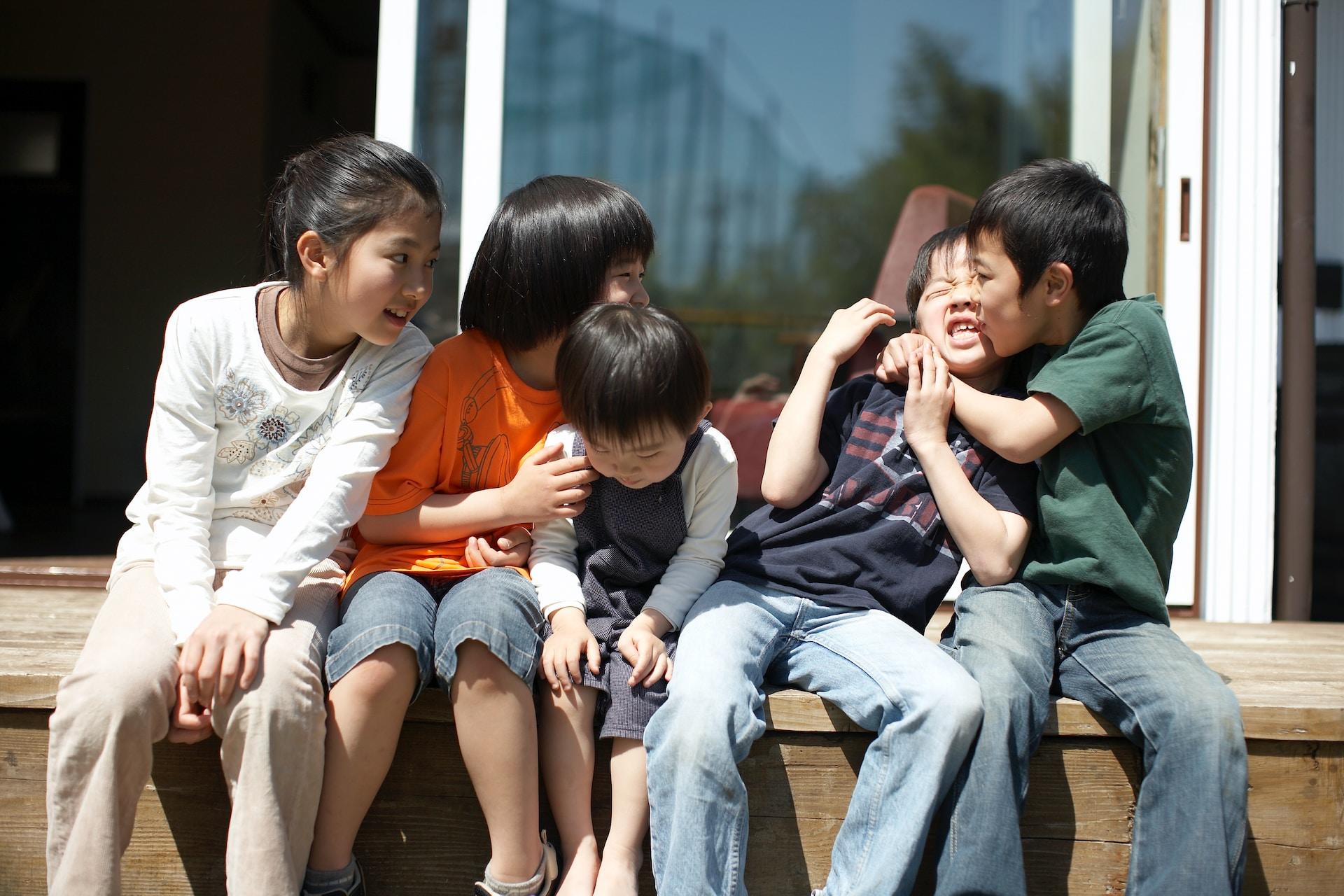 Five children of varying ages sit on a small ledge with the child on the furthest right holding the child next to him in a bear hug and kissing his cheek.