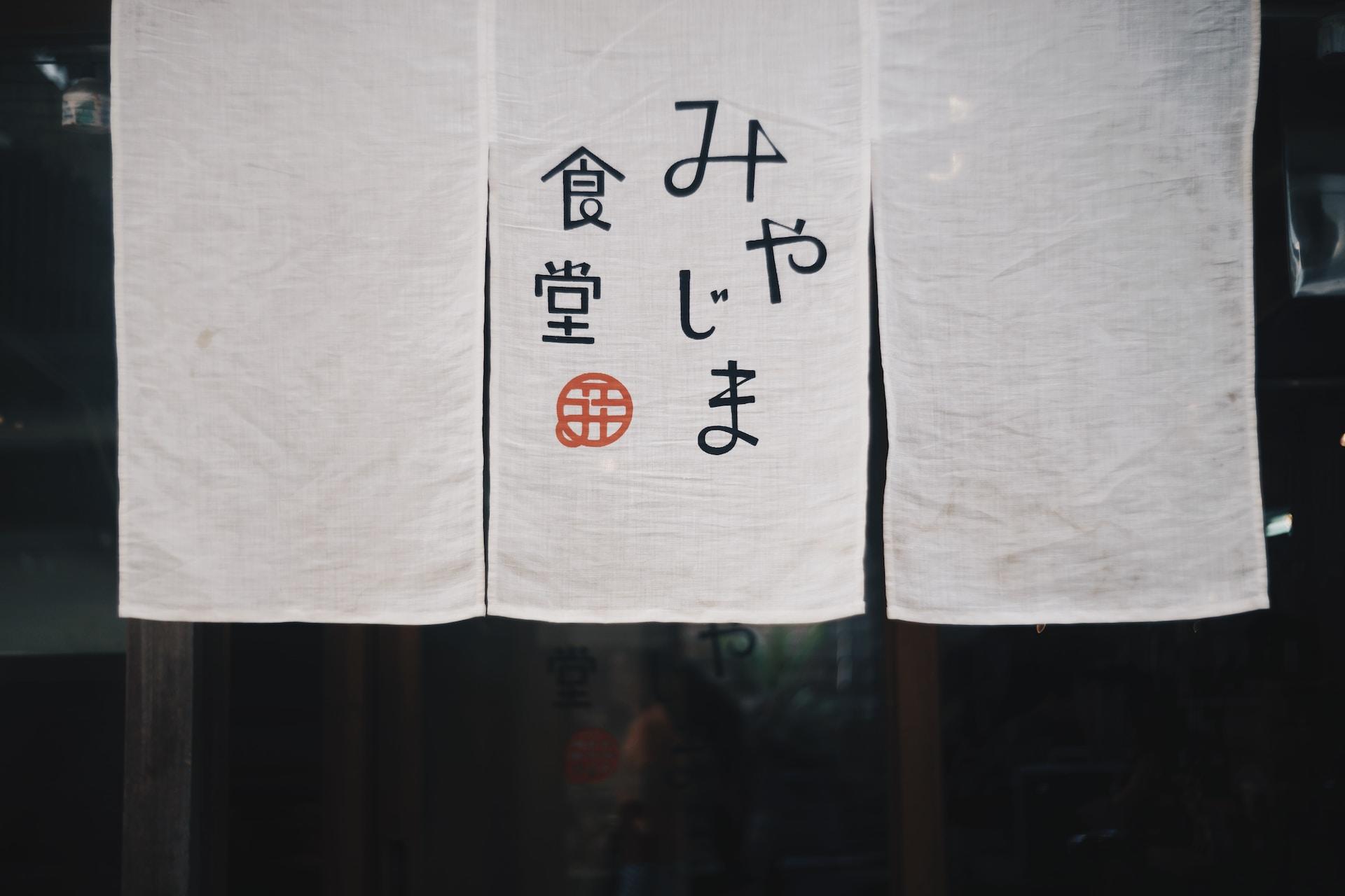 Three sheets of white cloth hung portrait-style from a rod with the middle one featuring Japanese writing in black ink and a red seal in the lower-left quadrant