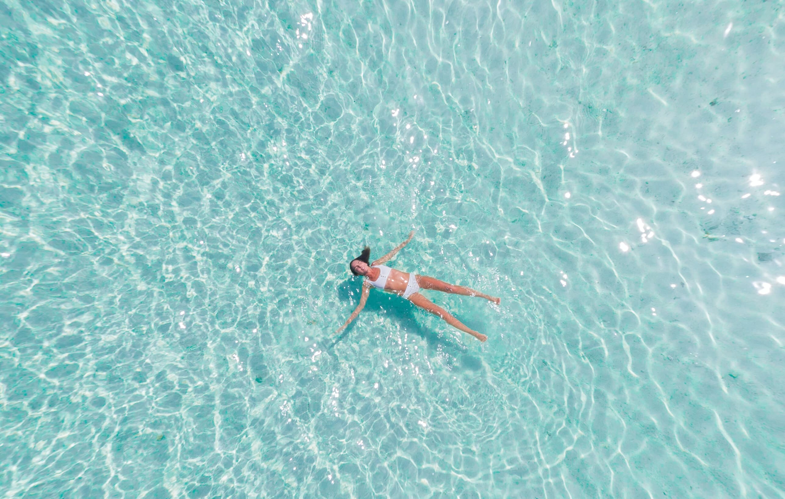a woman wearing a white two-piece swimsuit floats face-up in a greenish body of water with her arms and legs outstretched. 