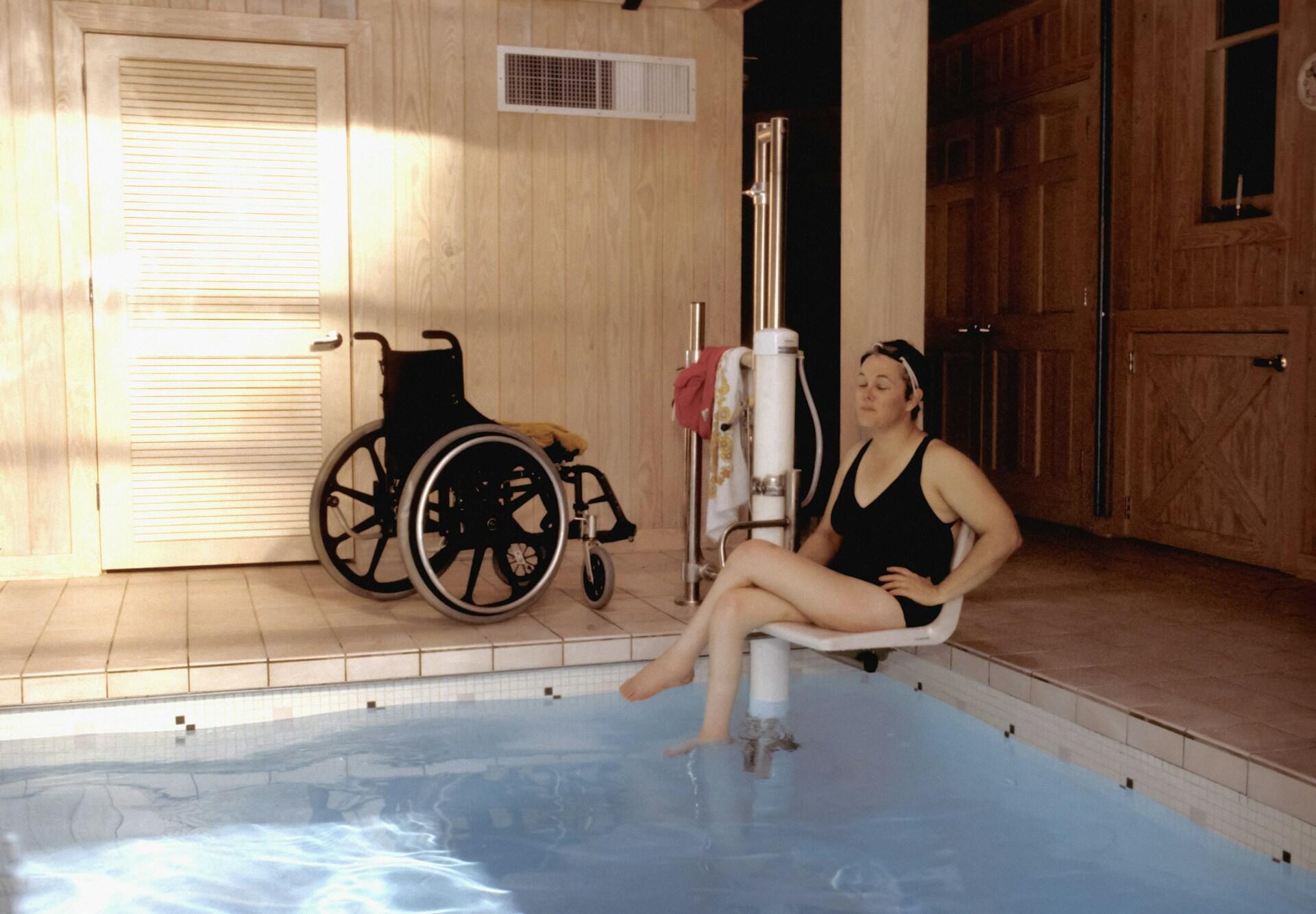 A woman in a black bathing suit sits on a pool lift with a wheelchair nearby.