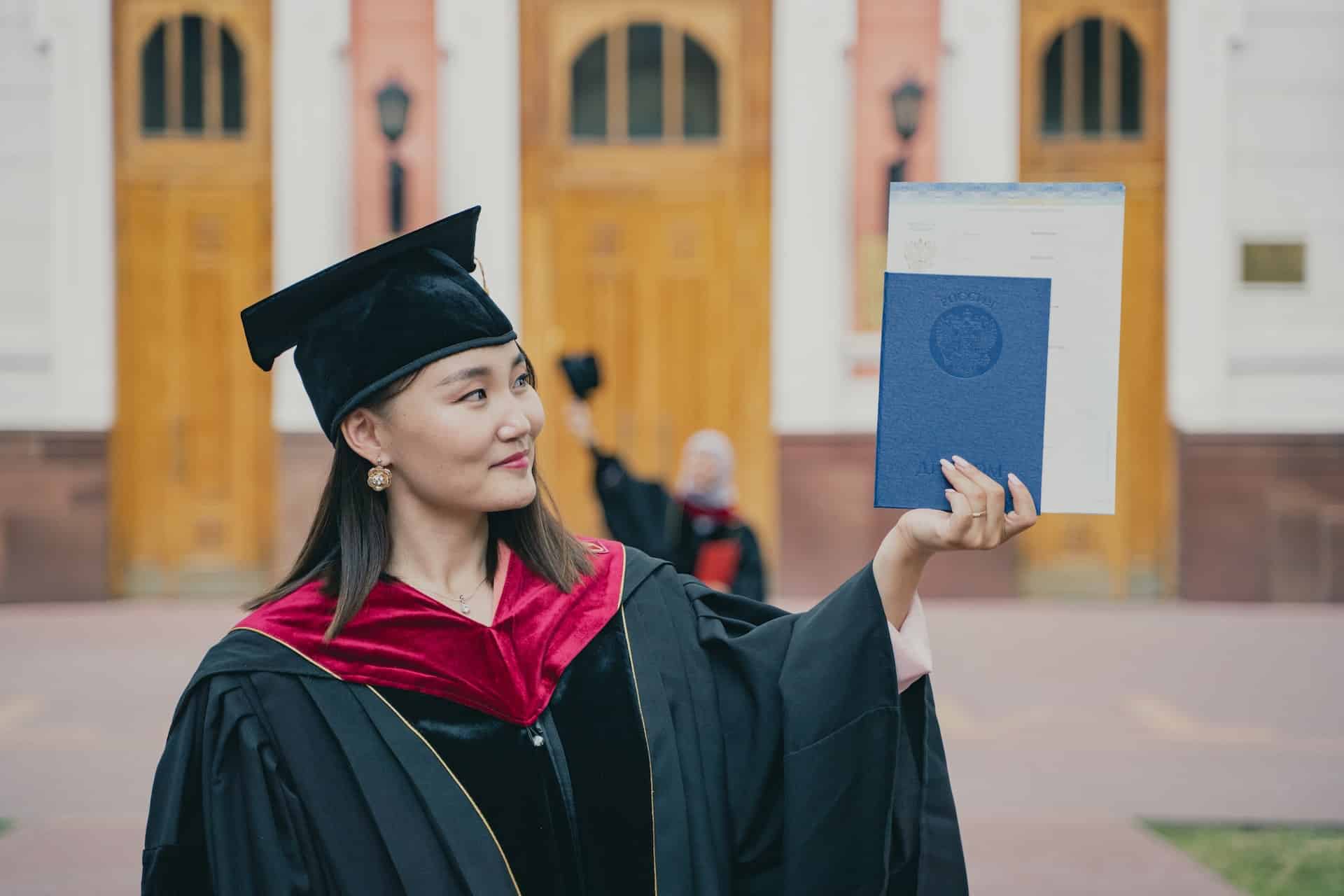 A woman in black graduation regalia with a dark red sash holds up her diploma