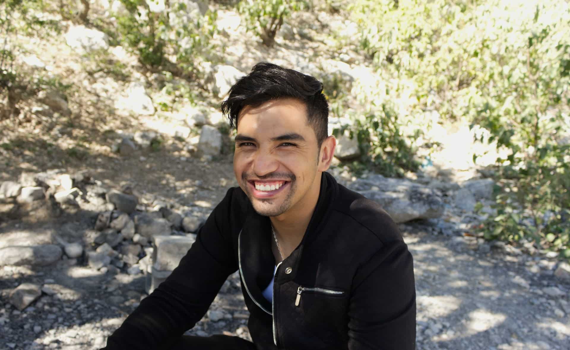 A dark-haired man in a black, zip-up shirt, sitting outdoors