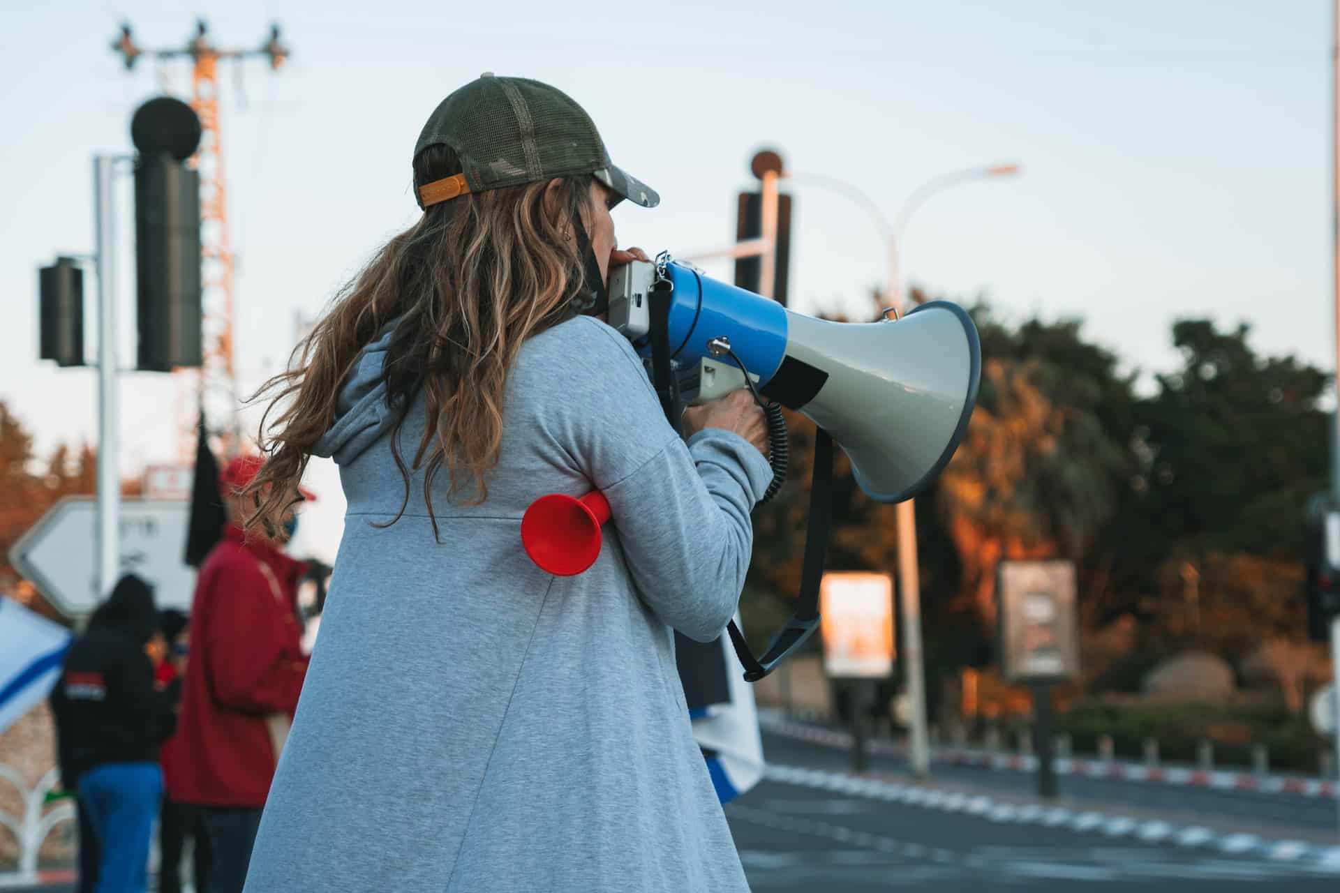 A woman with long hair, wearing wearing a light blue jacket and speaking into a bullhorn while standing on the street.