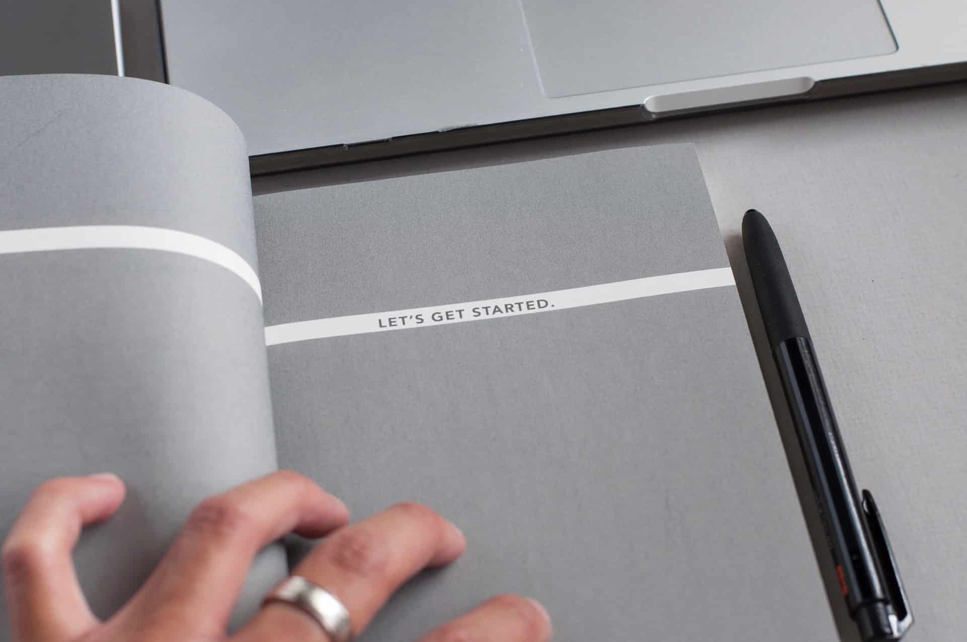 A black pen next to a grey notebook with 'let's get started' printed on a white ribbon.