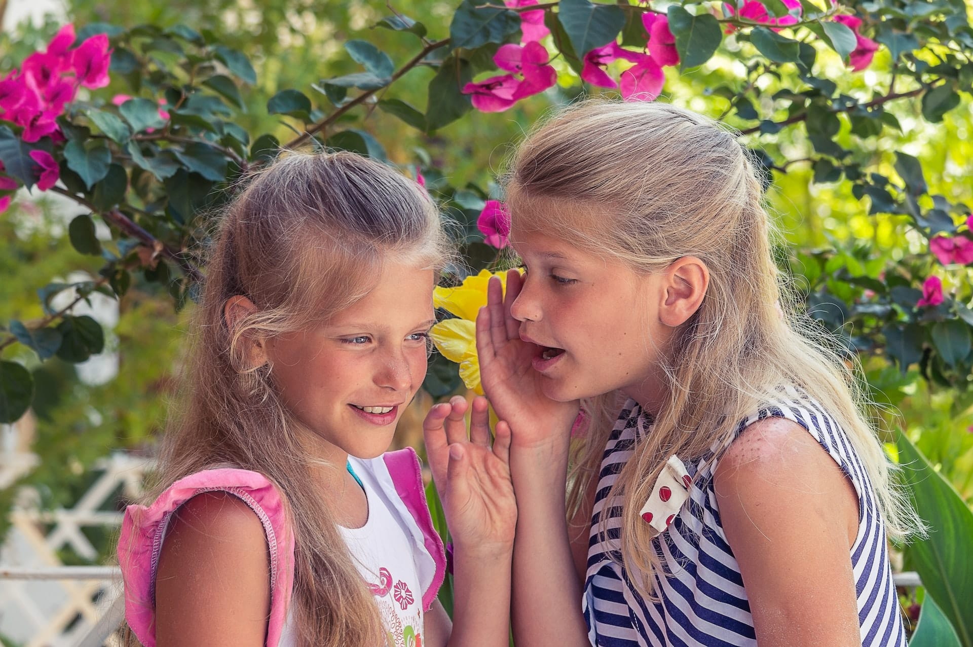 Two blond girls standing in a flowery garden, sharing a secret.