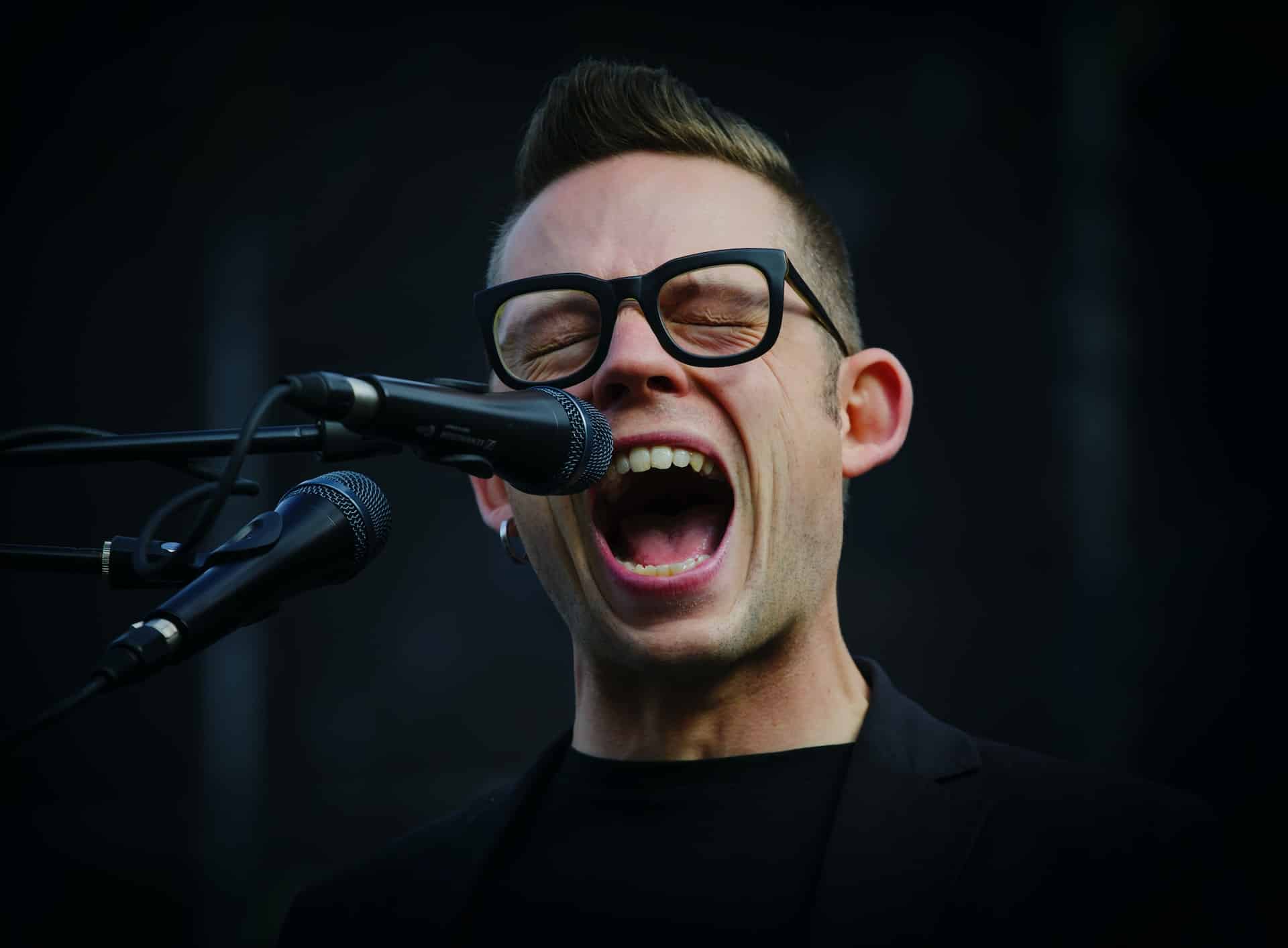 A man in a black t-shirt wearing dark-framed glasses sings into a microphone against a black background