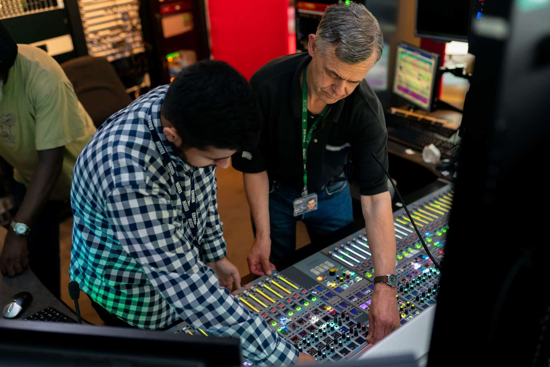 An older man in a black tee-shirt showing a younger trainee in a checkered shirt how to wire a music studio soundboard.