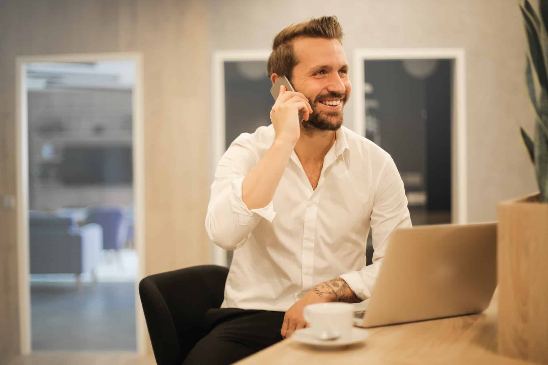 A bearded man in a white shirt smiling and talking on his mobile while seated at his desk.