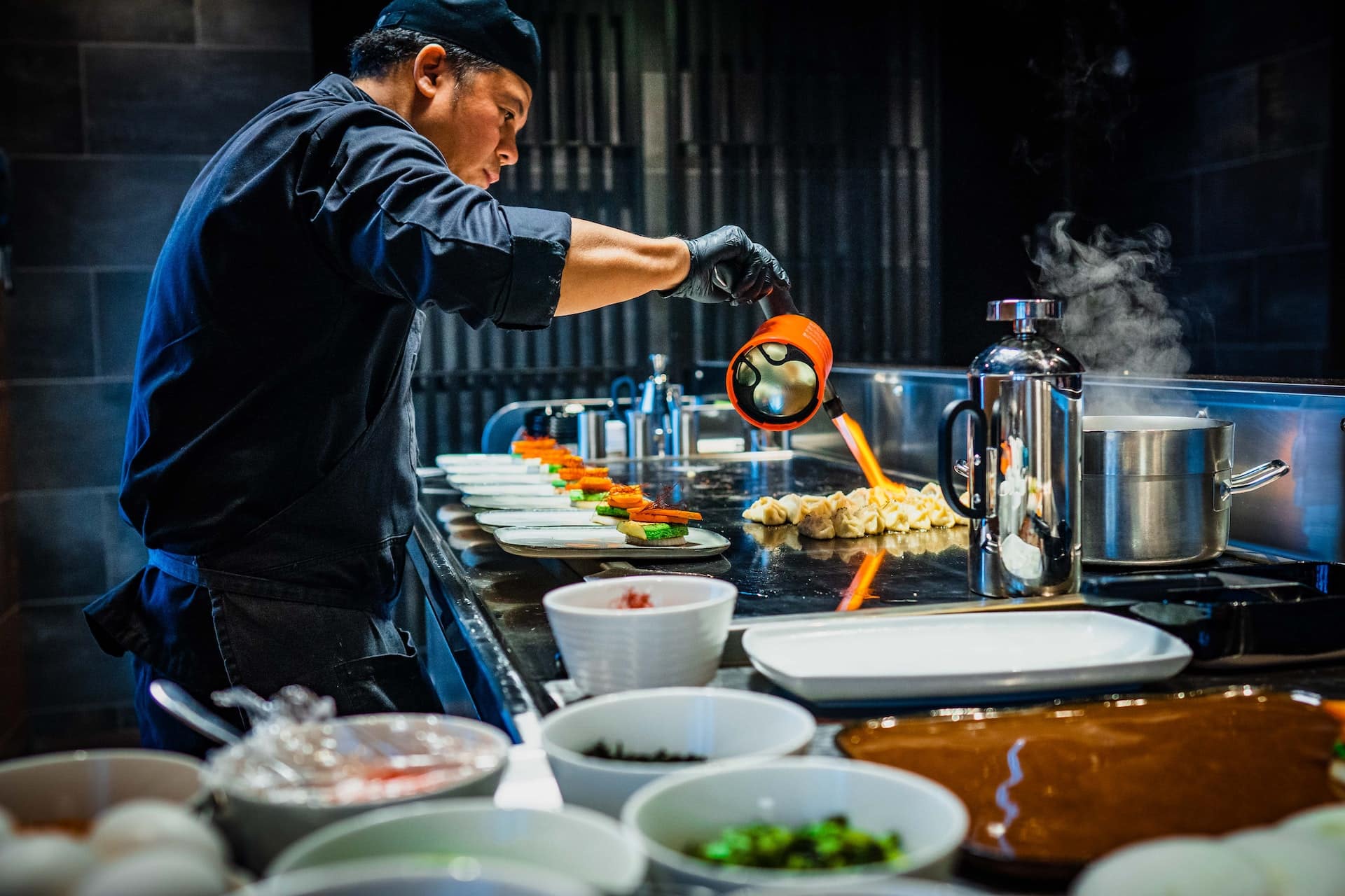 A black-clad chef in his kitchen wielding a pastry burner over some desserts.