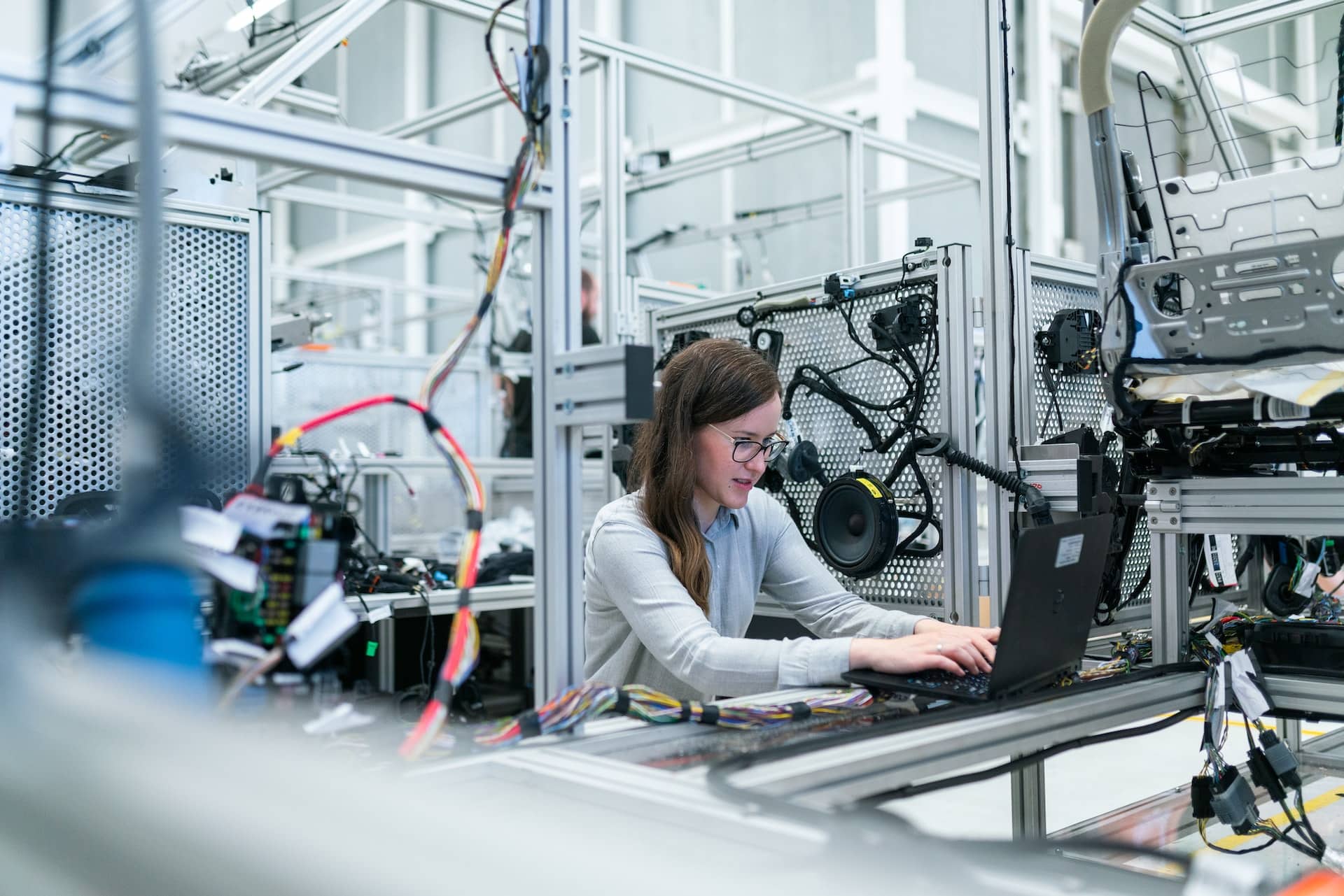 A dark-haired woman with glasses sitting and typing on a laptop inside a robotic programming cage