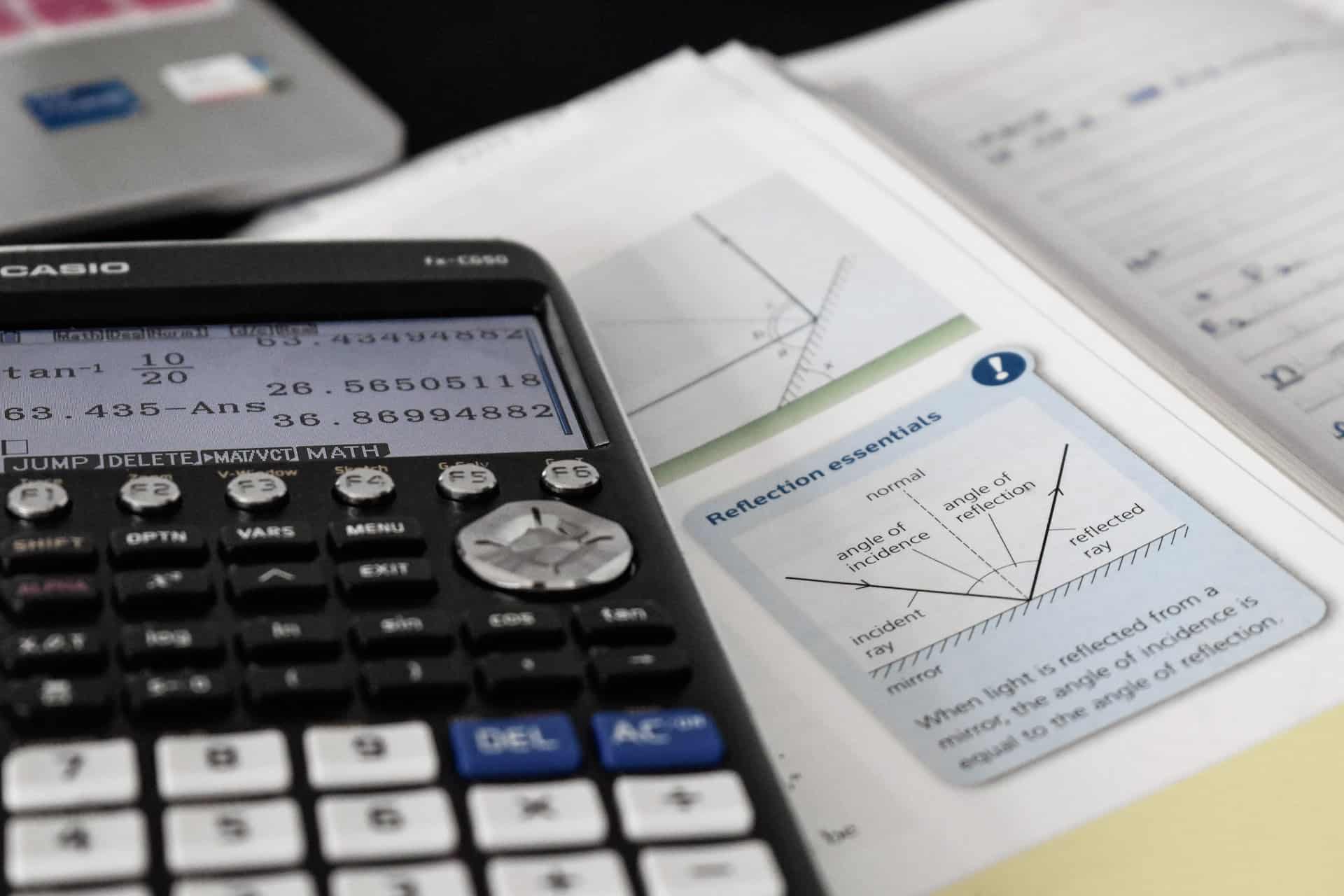 A black calculator with white numerical buttons displaying scientific notation, laying atop a booklet of maths problems