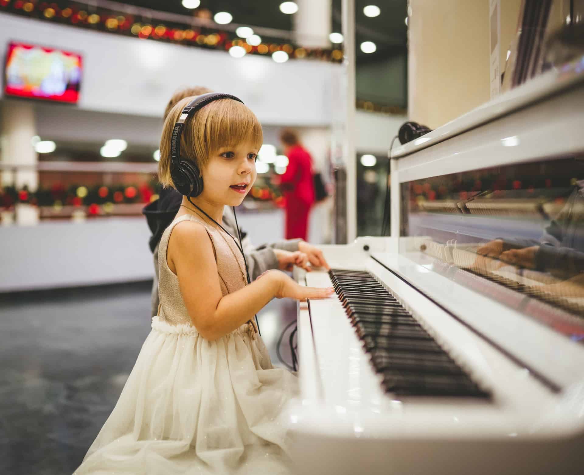 A small girl in a white dress with headphones on sits at the piano, ready to play