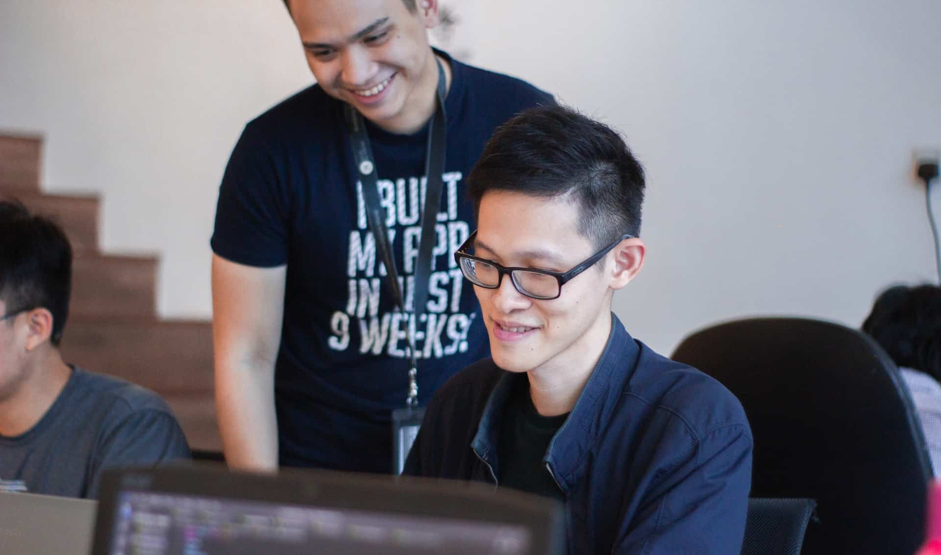 A dark-haired man in a black shirt sits and smiles in front of a computer while another dark-shirted man stands behind him, smiling and looking at the computer