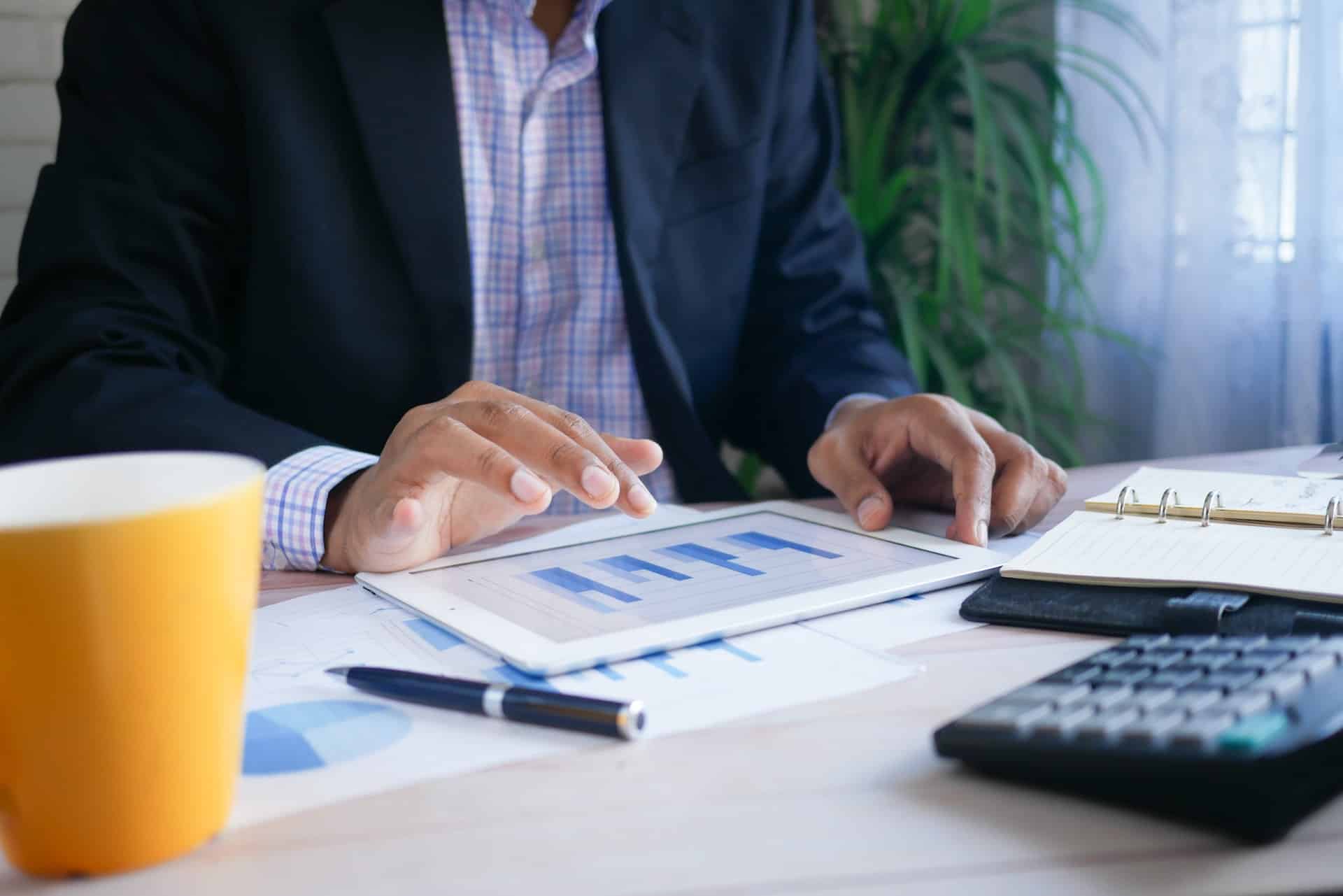 A man wearing a suit jacket over a plaid shirt looks at a blue-toned bar graph on a tablet; there's a yellow cup nearby