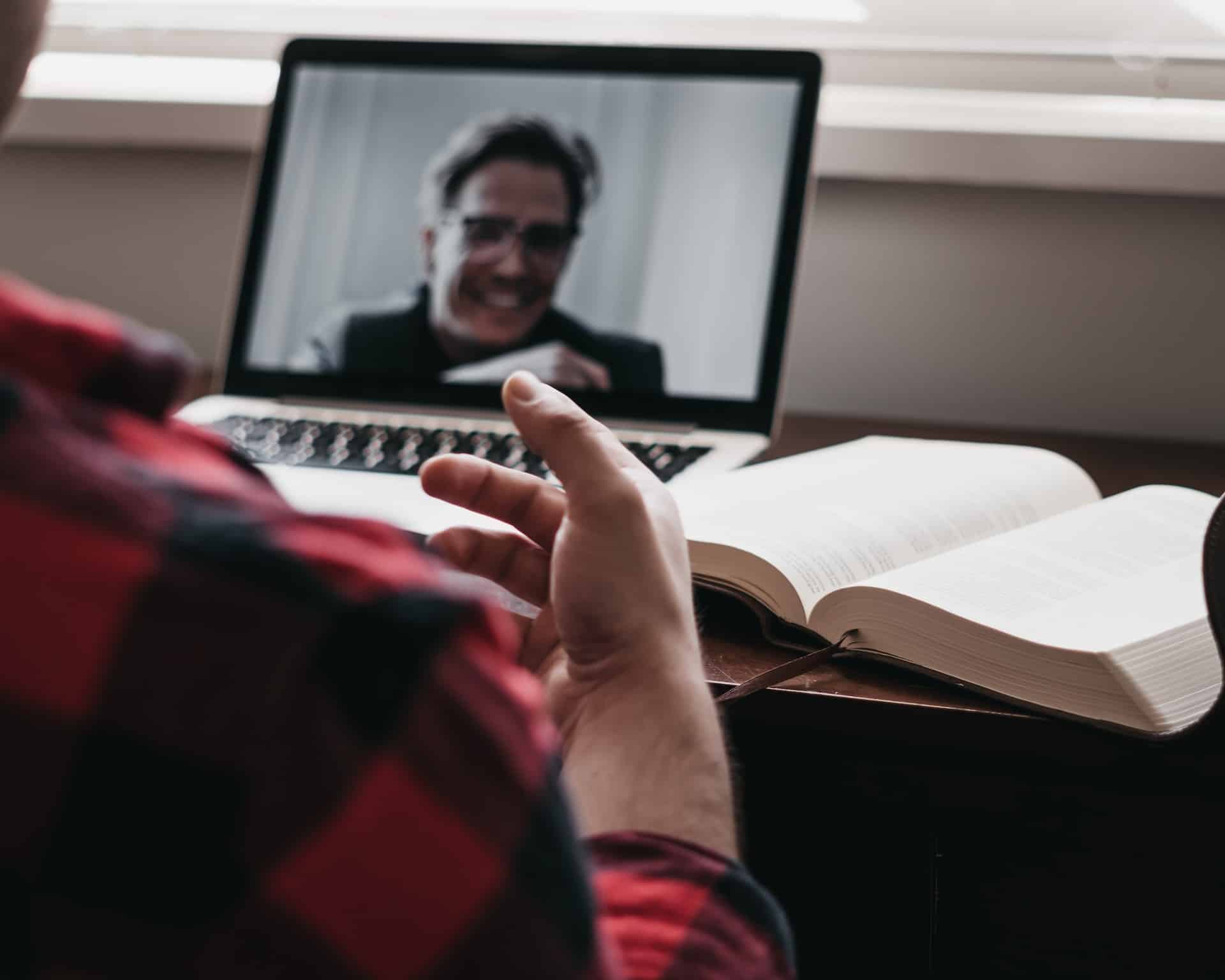 A person in a red and black plaid shirt sits at a table in front of an open book and open laptop computer, speaking to someone remotely