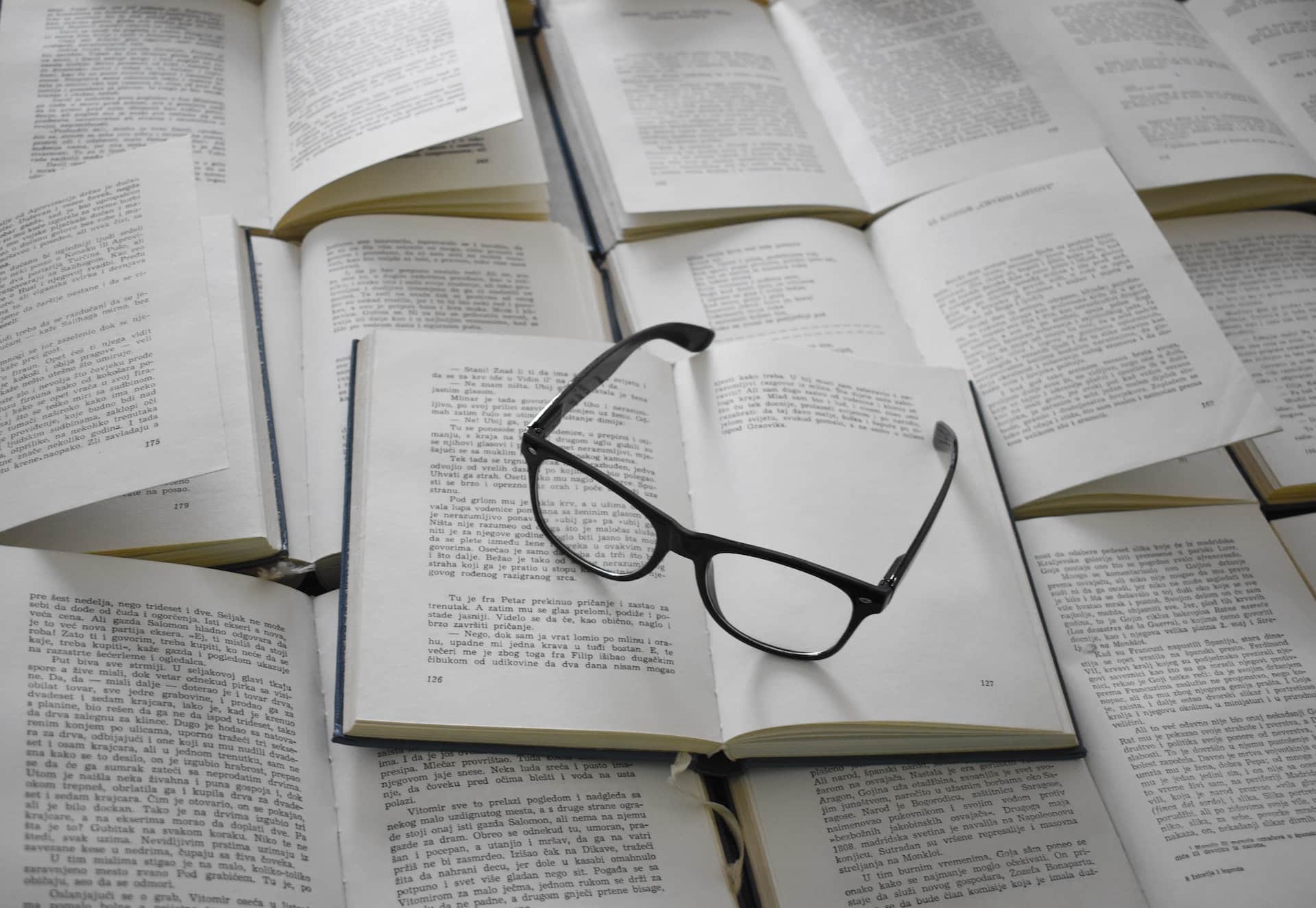 A scattering of open books with a pair of eyeglasses resting atop of them.