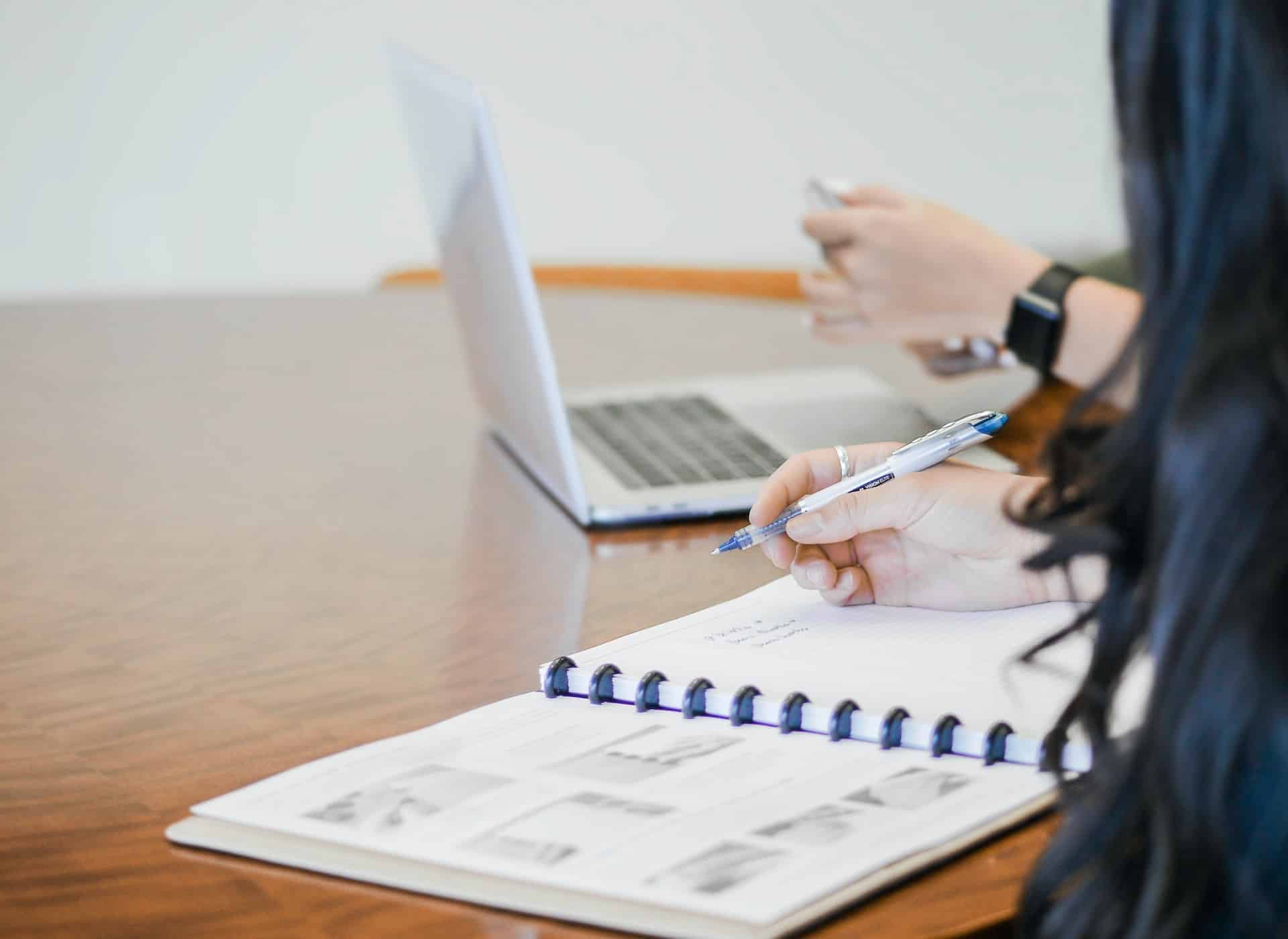 A woman with long, dark hair sits at a table in front of an open, spiral-bound notebook, holding a blue ink pen in her right hand