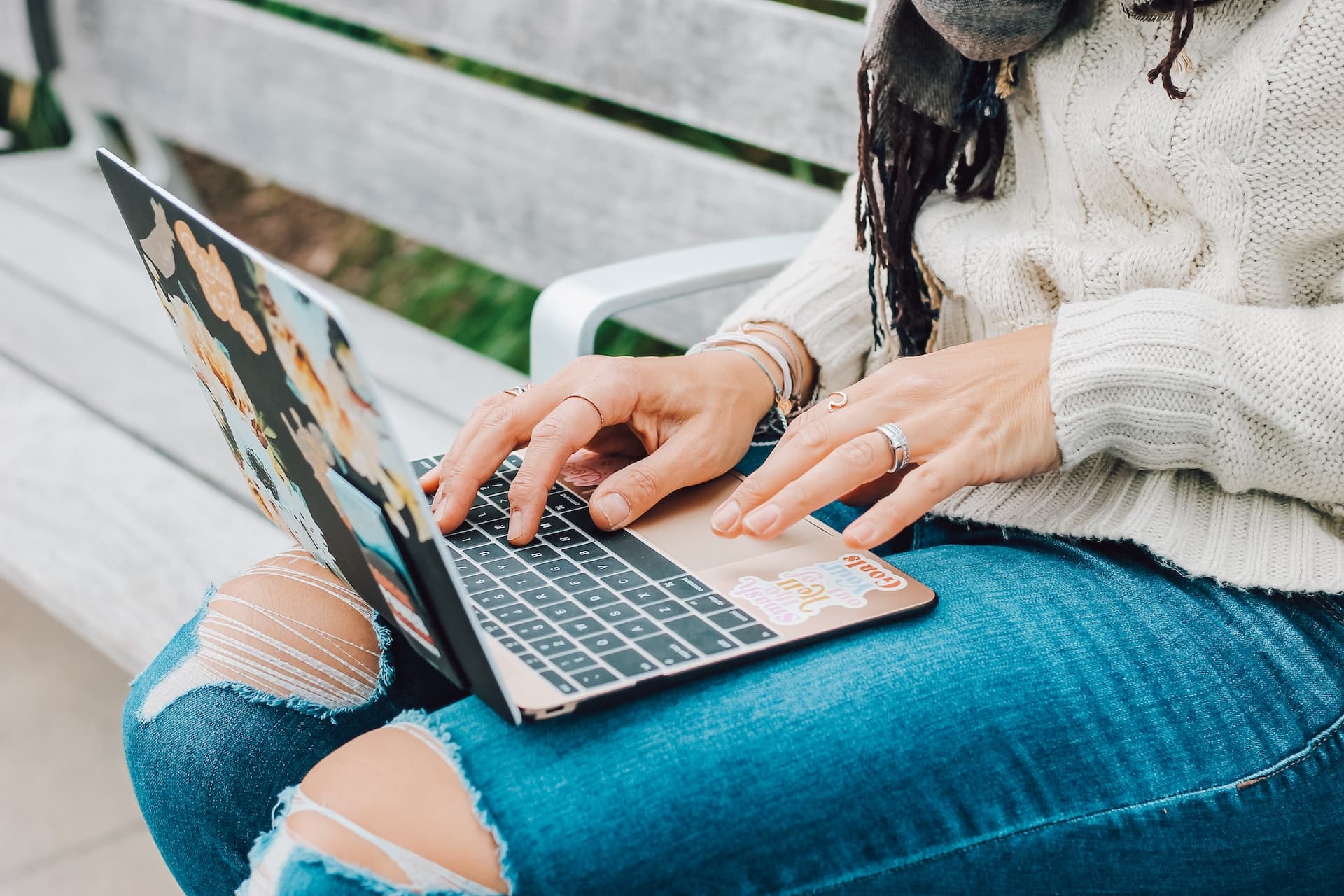 A woman in torn jeans and taupe jumper works on the open laptop propped on her lap