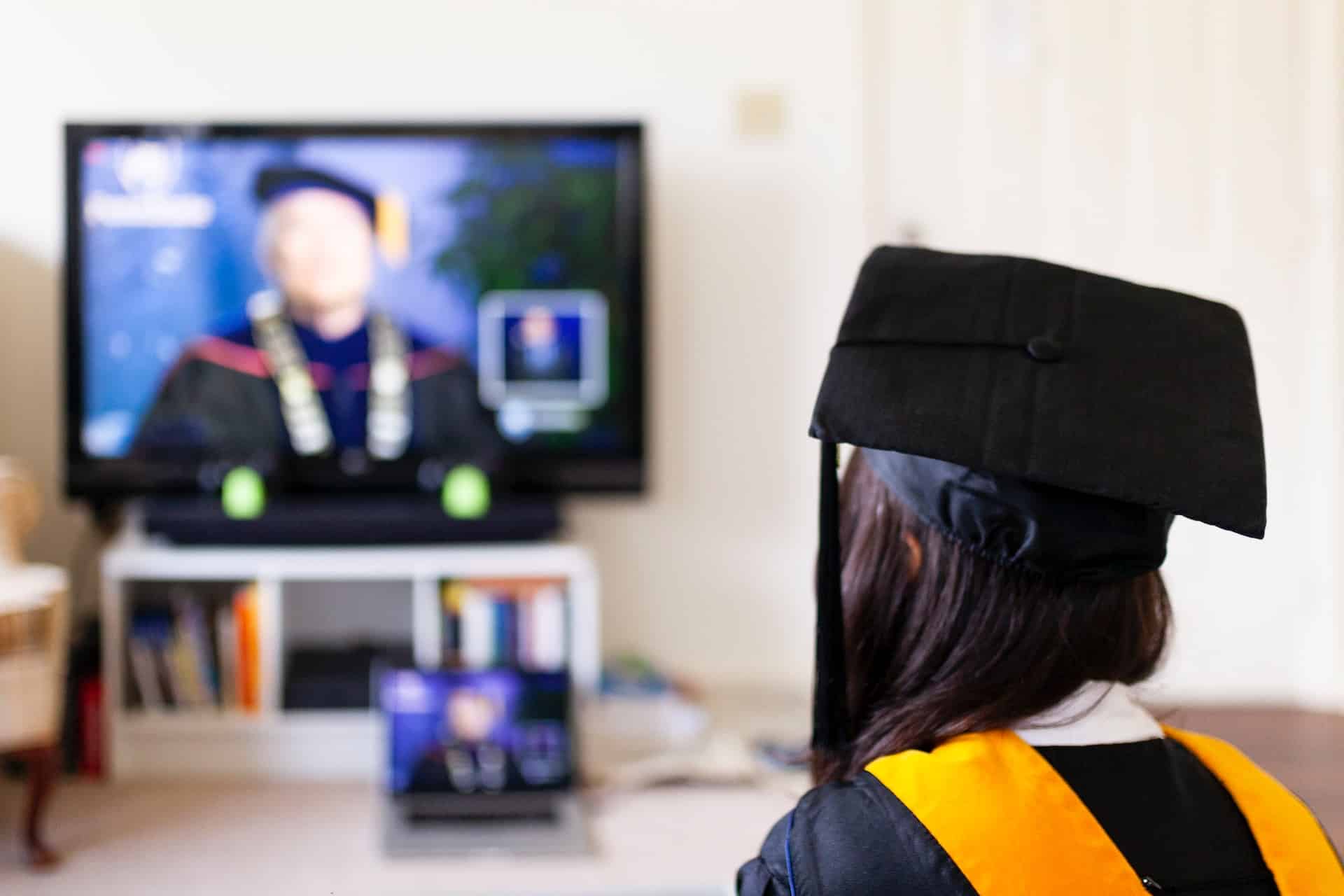 A student in graduation regalia sits in front of a computer monitor displaying a university professor in regalia