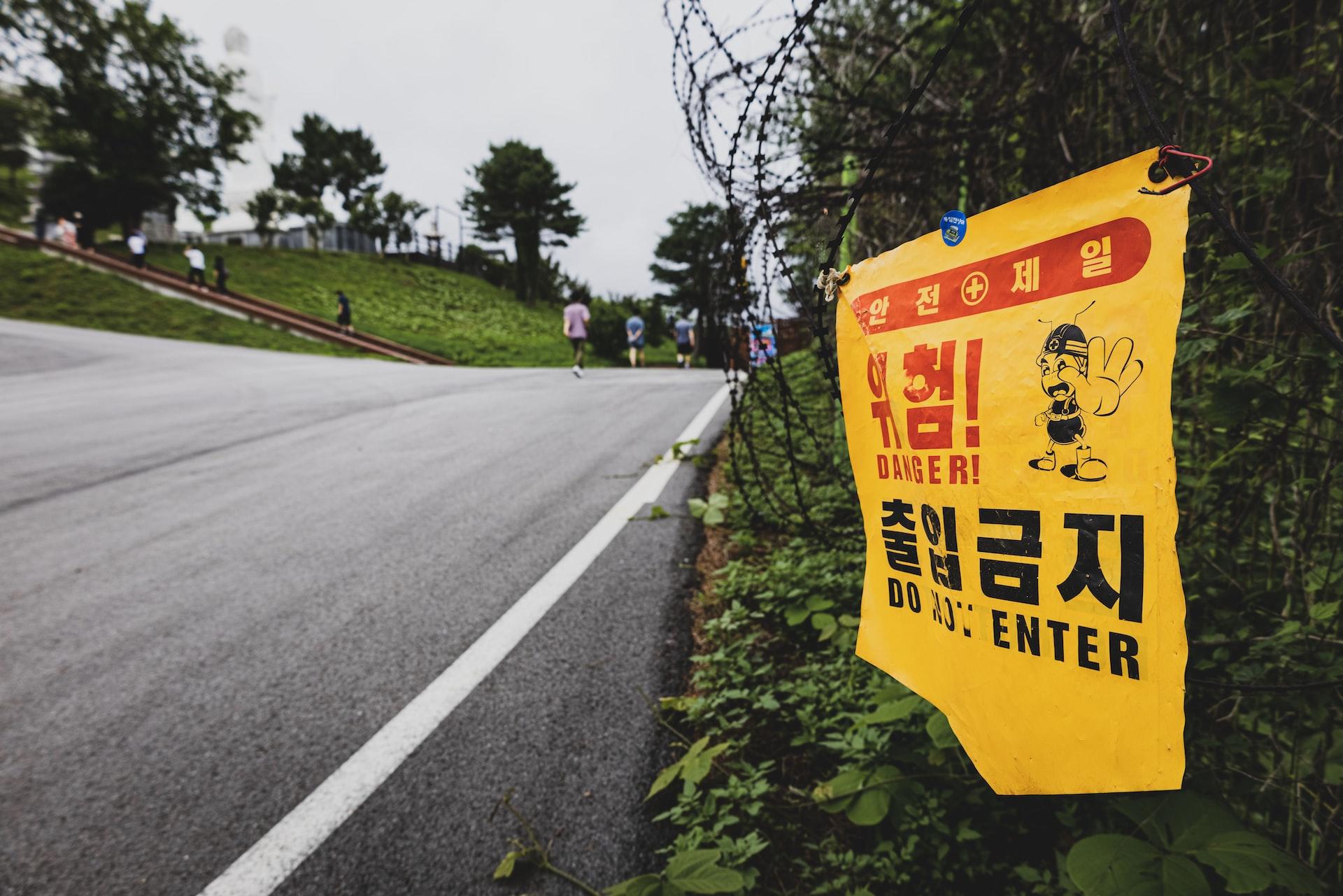 An empty road with a prominent white stripe along its side with a yellow sign warning of danger and people in the background, climbing a hill