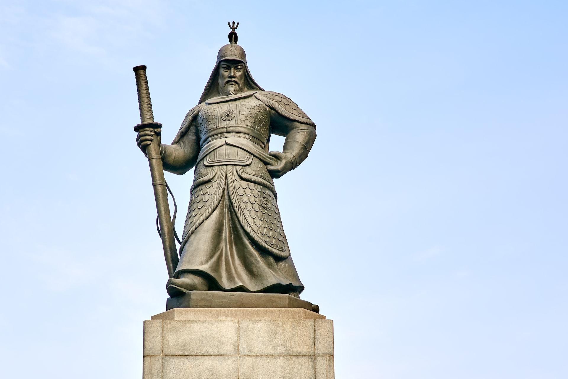 A sculpture of Yi Sun-sin, the Creator of the Turtle Ship, mounted on a concrete pedestal, set against a pale blue sky.