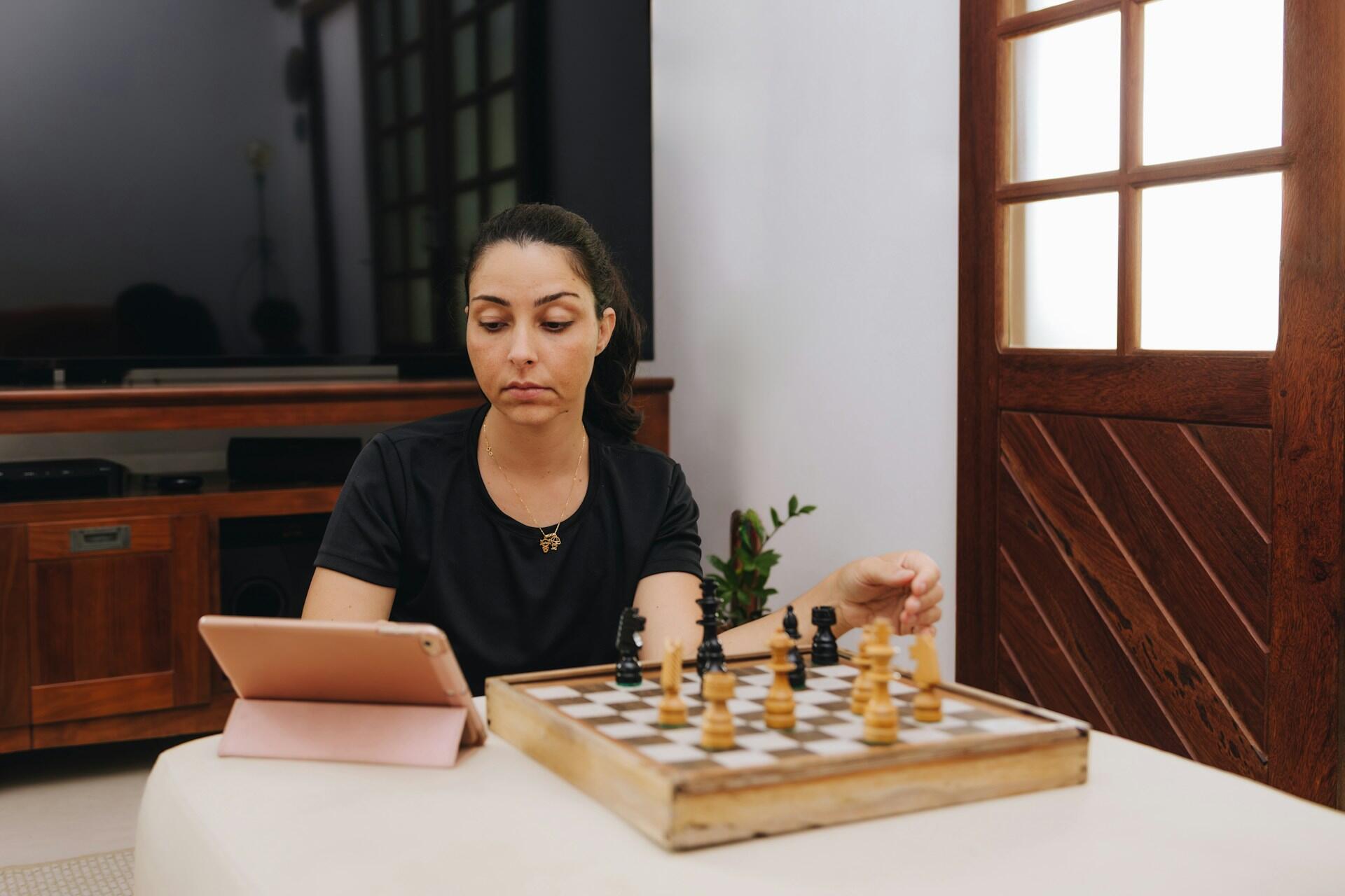 A person in a black teeshirt sitting in front of a chessboard with a tablet propped up beside it.
