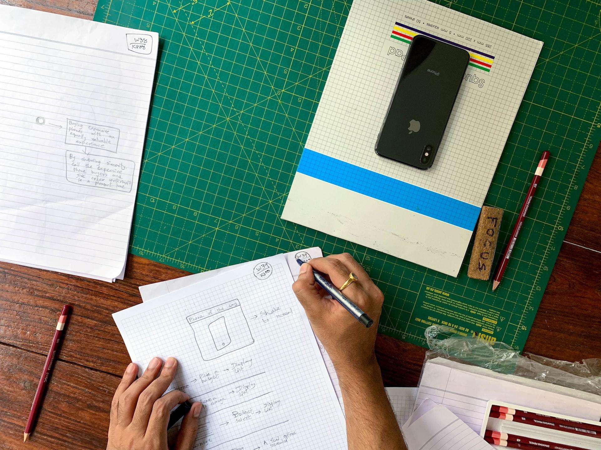 A person takes notes on a table crowded with a green grid board, a notebook, a rubber and two red pencils. 
