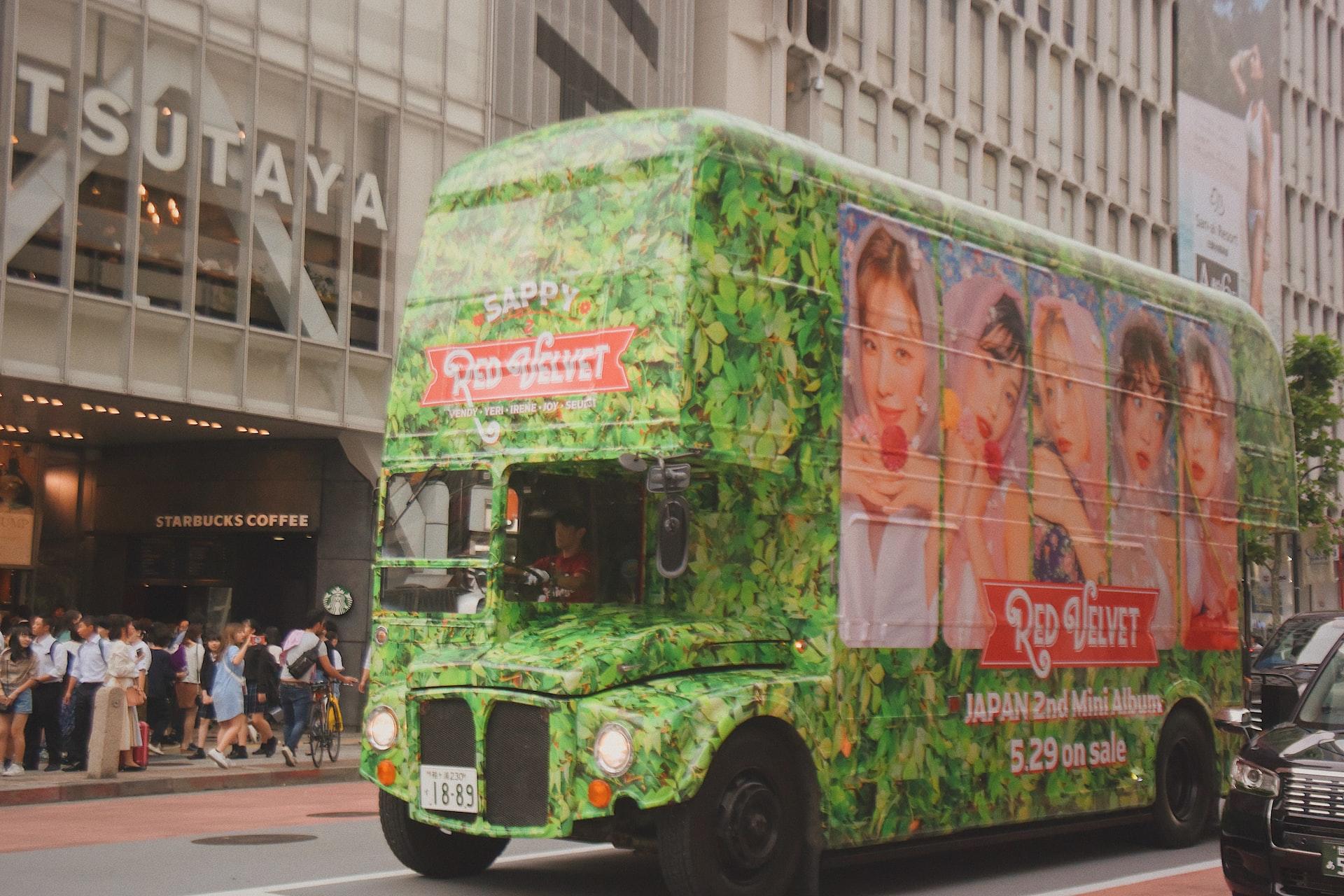 A green double-decker bus with an advert for the K-pop group Red Velvet displayed on it riding down a city street in Japan.