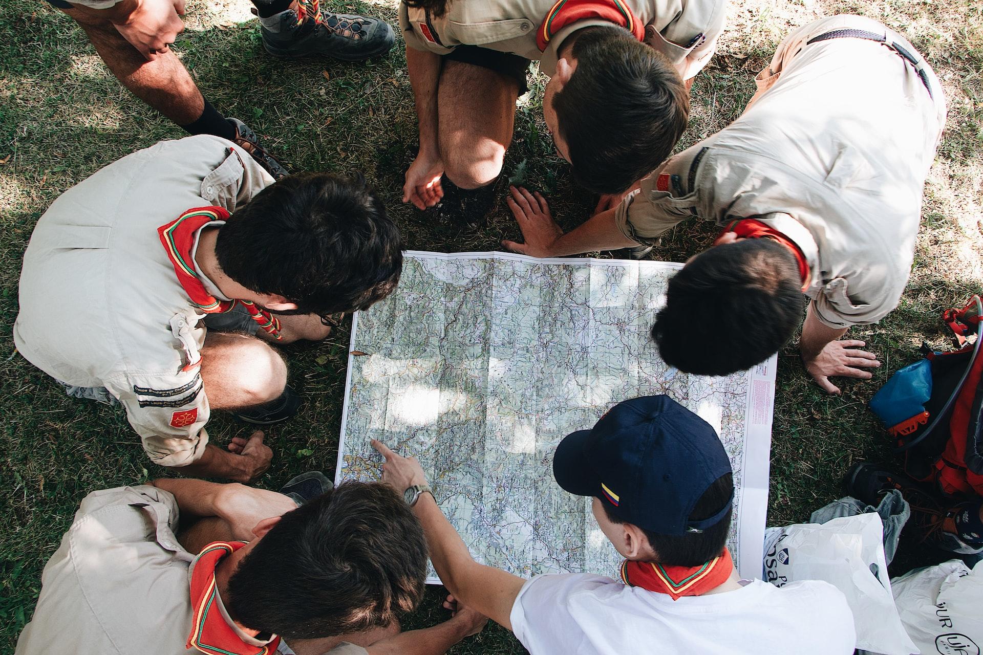 A group of boys with black hair, wearing scouts' uniforms, kneel on the ground to study a map.